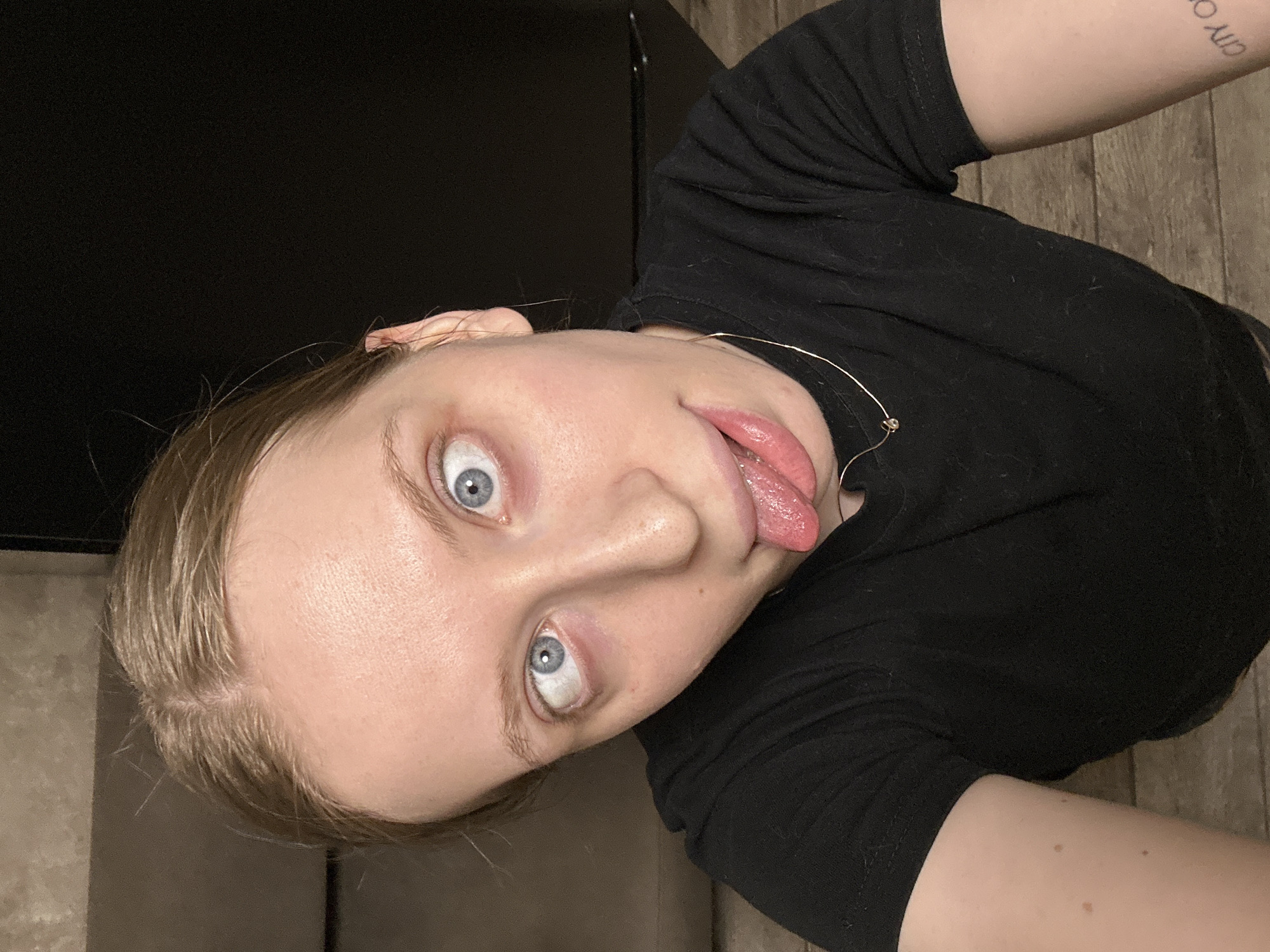 A close-up, rotated portrait shot features a young individual, likely female, with light blonde or light brown hair and striking blue eyes. The subject's head is tilted dramatically, with their face on the left side of the frame, and their eyes are wide open, directly engaging the camera. A playful or silly expression is conveyed by their tongue extended outwards from their mouth.
The person wears a black, short-sleeved t-shirt and a thin, delicate gold-colored necklace. A partial tattoo is visible on their upper right arm, showing text that appears to begin with "CITY O". The skin on their arm also displays several small moles or freckles.
The background on the upper left of the image is dark and indistinct, possibly a wall or piece of furniture. On the lower right, a light brown wooden floor with visible grain serves as the backdrop. The lighting appears to be even, possibly from an indoor source, and the overall atmosphere is casual and expressive. The image is taken in Chaikovsky, Russia.