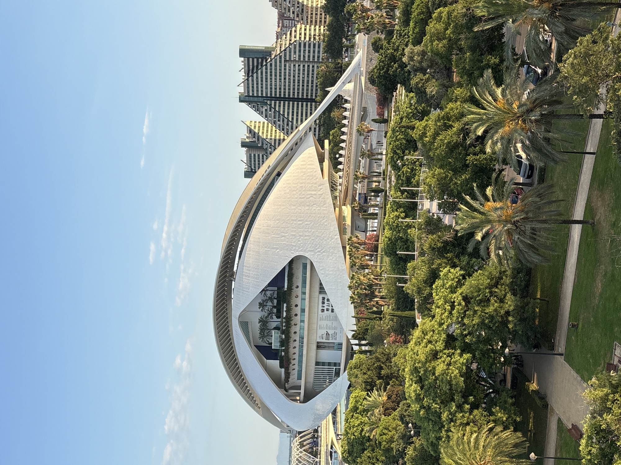 This media file showcases the striking architecture of the Hemisfèric, part of the City of Arts and Sciences in Valencia, Spain. The building, with its iconic eye-shaped design, dominates the frame, its white, futuristic structure contrasting with the lush green trees and palm trees surrounding it. The clear blue sky with scattered white clouds suggests it is a bright, sunny day, likely in the morning or late afternoon, given the gentle light.
The scene depicts a public space, with paved walkways and manicured lawns indicating a park-like setting. While no people are prominently featured, the presence of benches and lampposts suggests a place for recreation and leisurely strolls. In the background, modern buildings with unique geometric designs rise above the trees, adding to the contemporary architectural landscape of the area. Some cars are visible parked along a street or pathway, hinting at accessibility.
A closer look at the Hemisfèric reveals interior spaces through its open design, with what appears to be a pool or water feature visible inside. Text is visible on a poster or sign within the building, with the words "LES ARTS" and other French text legible, possibly indicating an exhibition or event. The overall impression is one of modern design, public enjoyment of an urban green space, and a visually appealing architectural landmark.