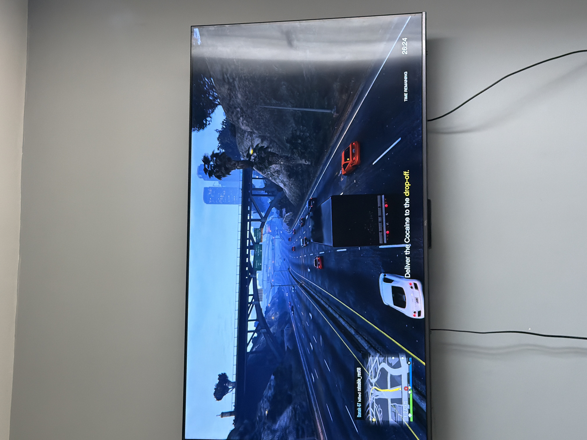 A television screen, likely a TCL brand model, is prominently displayed, mounted against a plain light gray wall in Dabene, Bulgaria. The screen is oriented vertically in the image, showing a video game in progress. The game appears to be an open-world driving simulation, possibly Grand Theft Auto V, depicted at dusk or night with a dark blue sky and illuminated cityscapes in the distance.
The game scene features a multi-lane highway or freeway, with several vehicles visible. An orange sports car is on the right side of the road, while a large black truck, likely the player's current vehicle, is in the center lanes. A white car follows behind the truck. The road is bordered by mountainous terrain with sparse vegetation, including palm trees, and is traversed by an overhead bridge structure.
The game's user interface is visible, indicating an active mission. A prominent yellow objective reads, "Deliver the Cocaine to the drop-off." A "TIME REMAINING" counter shows "26:24." In the bottom left, a mini-map displays the current location, marked route, and a distance of "12.48km." A kill notification states, "Danak-67 killed rateable_rest18."
A black cable runs along the light gray wall to the right of the television screen. The lighting in the room suggests an indoor setting, with ambient light reflecting off the screen surface.