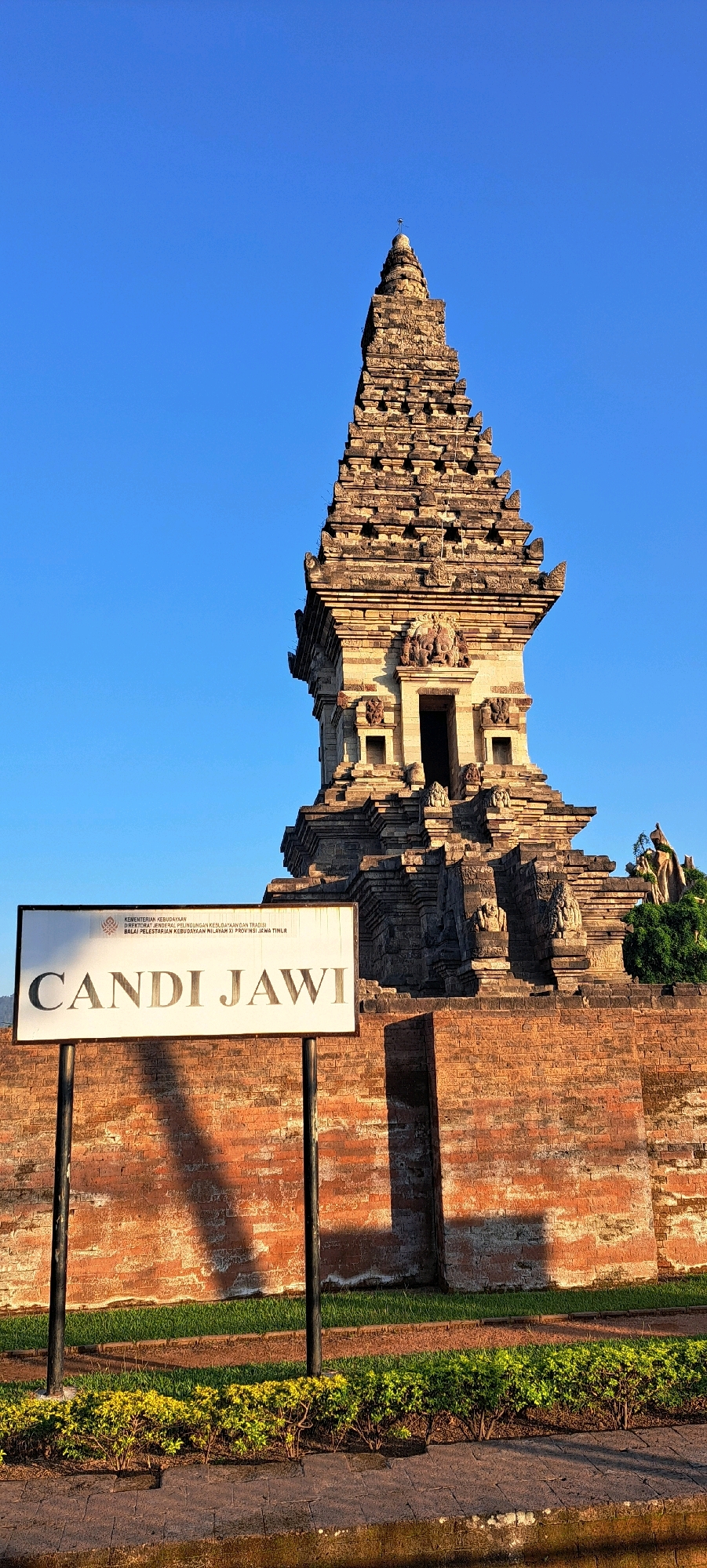 The image shows a temple, identified by a sign in the foreground as "Candi Jawi," located in Prigen, Indonesia. The temple is made of stone and has a tiered, pyramidal structure. A sign in front of the temple reads "KEMENTERIAN KEBUDAYAAN, KESENIAN DAN WARISAN" and "BALAI PELESTARIAN KEBUDAYAAN WILAYAH XI PROVINSI JAWA TIMUR" above the name of the temple. The temple is situated behind a brick wall, with a strip of grass and low-lying bushes in front of the wall. The sky is clear and blue, suggesting a daytime scene, possibly in the late afternoon given the long shadows cast on the wall and ground. There are no people visible in the image. The architecture of the temple suggests a historical or cultural site.