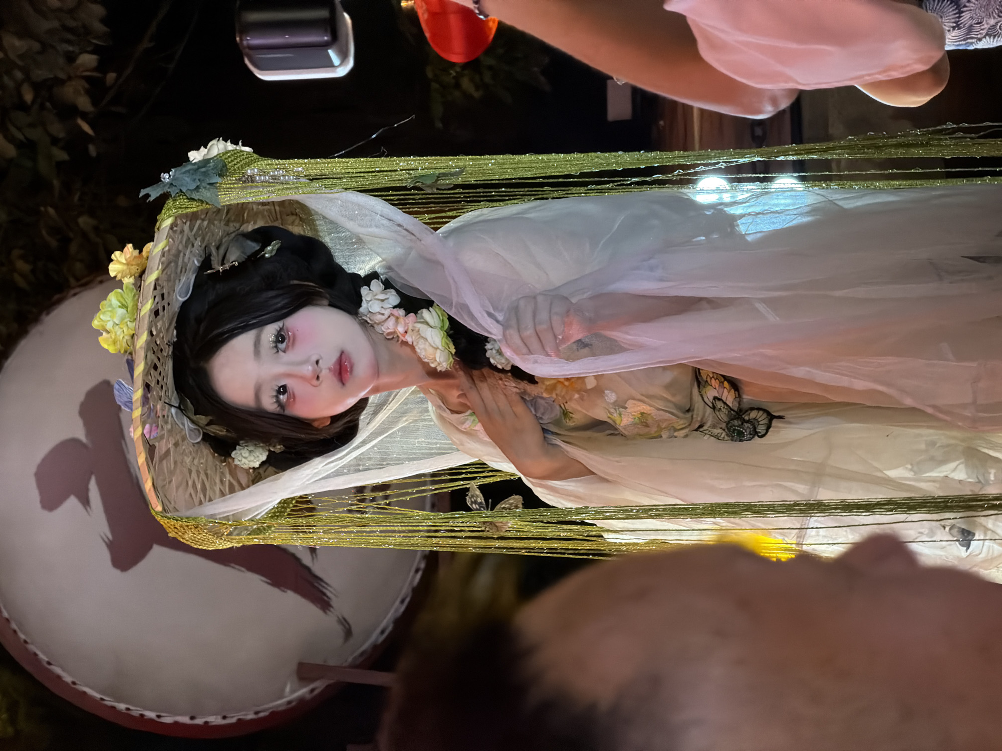 In Yuzhong District, China, a young woman adorned in a traditional Chinese costume stands prominently. Her attire consists of a layered robe featuring floral embroidery and a dramatic butterfly motif. She wears an elaborate straw hat, decorated with fabric flowers and delicate netting that drapes around her face. Her makeup is striking, with exaggerated blush and glittery eyeshadow, emphasizing a theatrical presentation. She holds one hand to her chest, her other hand gently touching the sheer fabric cascading from her hat. The backdrop suggests an outdoor night setting, with a large, partially visible drum to her left, bearing a large Chinese character. Other elements of the scene include blurred figures in the foreground and background, a red lantern, and lighting that casts a soft glow on the subjects.