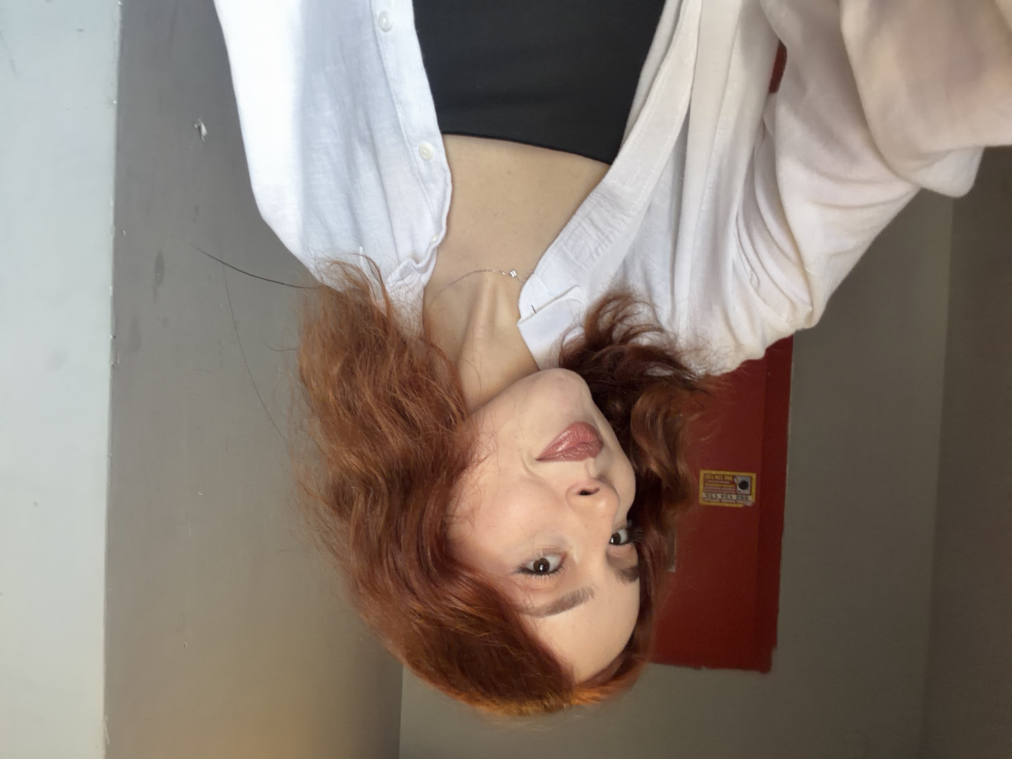 An eye-level, upright shot captures a young woman with reddish-brown, wavy hair looking directly at the camera with a subtle, calm expression. She has fair skin, light-colored eyes, and full lips adorned with a glossy, natural-toned lipstick. Her eyebrows are well-defined.
She is dressed in a white, unbuttoned collared shirt, worn over a black top that is partially visible underneath, possibly a crop top or camisole. A delicate silver-toned necklace with a small, square pendant is visible around her neck, resting on her collarbone.
The scene is an indoor setting with plain walls. To the left of the frame, a corner of a light-colored, possibly white, wall is visible. Behind her, to the right, there is a prominent red panel or door, which appears to be a utility fixture. A small, faded yellow sticker is affixed to this red object. The text on the sticker includes "SAEKUL" or "SEKLE" at the top and a series of numbers, resembling "875 512 000," below it, suggesting a phone number or identification code. The wall adjacent to the red panel is a neutral greyish-brown color. The lighting suggests it is daytime indoors, likely under natural or soft artificial light. The location context provided is Tbilisi, Georgia.