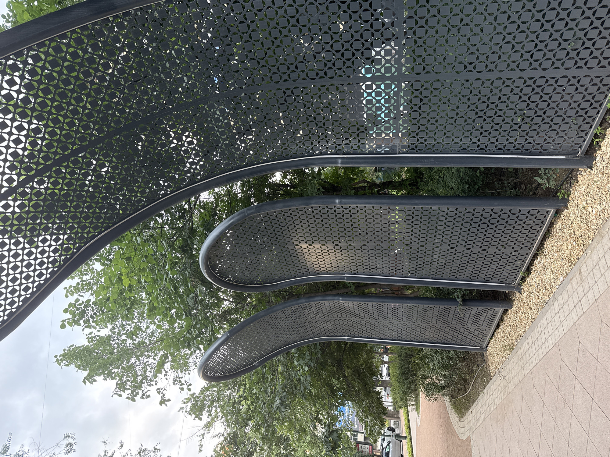 The image shows a section of a modern, curved, dark gray metal screen structure with an intricate geometric pattern. Three distinct curved panels are visible, forming an arched walkway or barrier. The panels are perforated with a repeating pattern of interconnected squares and circles. Green foliage, likely trees and bushes, is visible behind the screens, suggesting an outdoor urban setting. In the background, there are buildings and cars, indicating a city environment. The ground in the foreground is paved with light brown tiles and bordered by a path of small, light-colored pebbles. The sky is overcast with clouds. The image appears to be taken in Almaty, Kazakhstan, likely in a public space such as a park, plaza, or a street with modern landscaping. No people are visible in the frame.