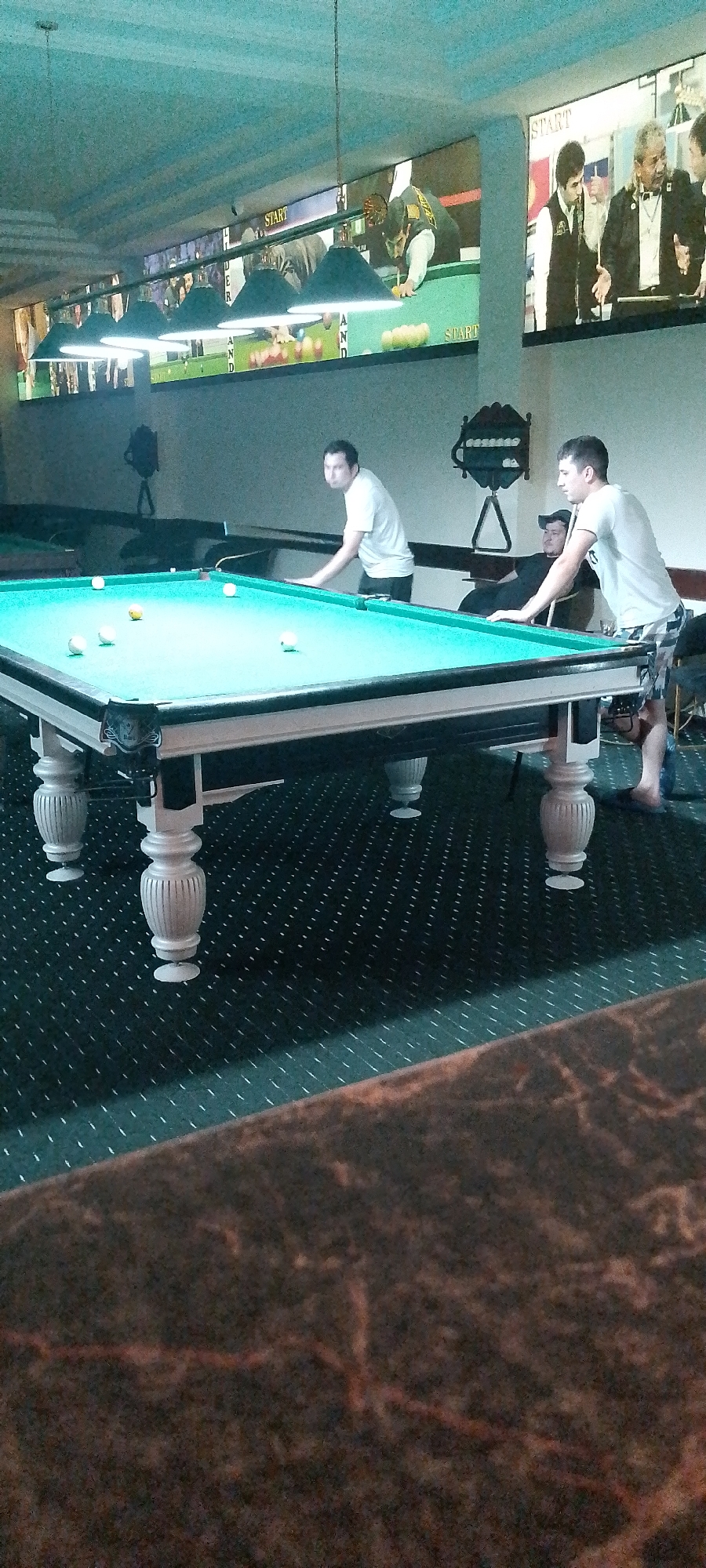 A billiard hall in Angren, Uzbekistan features a pool table with green felt and white cushions. Several billiard balls are scattered on the table. Two men are playing pool, one in a white t-shirt and black shorts, the other in a white t-shirt and patterned shorts. A third man in a black cap is seated in the background. Above the pool table, a large screen displays a video of people playing billiards. The room has a dimly lit, teal-colored ceiling and patterned dark green carpet.