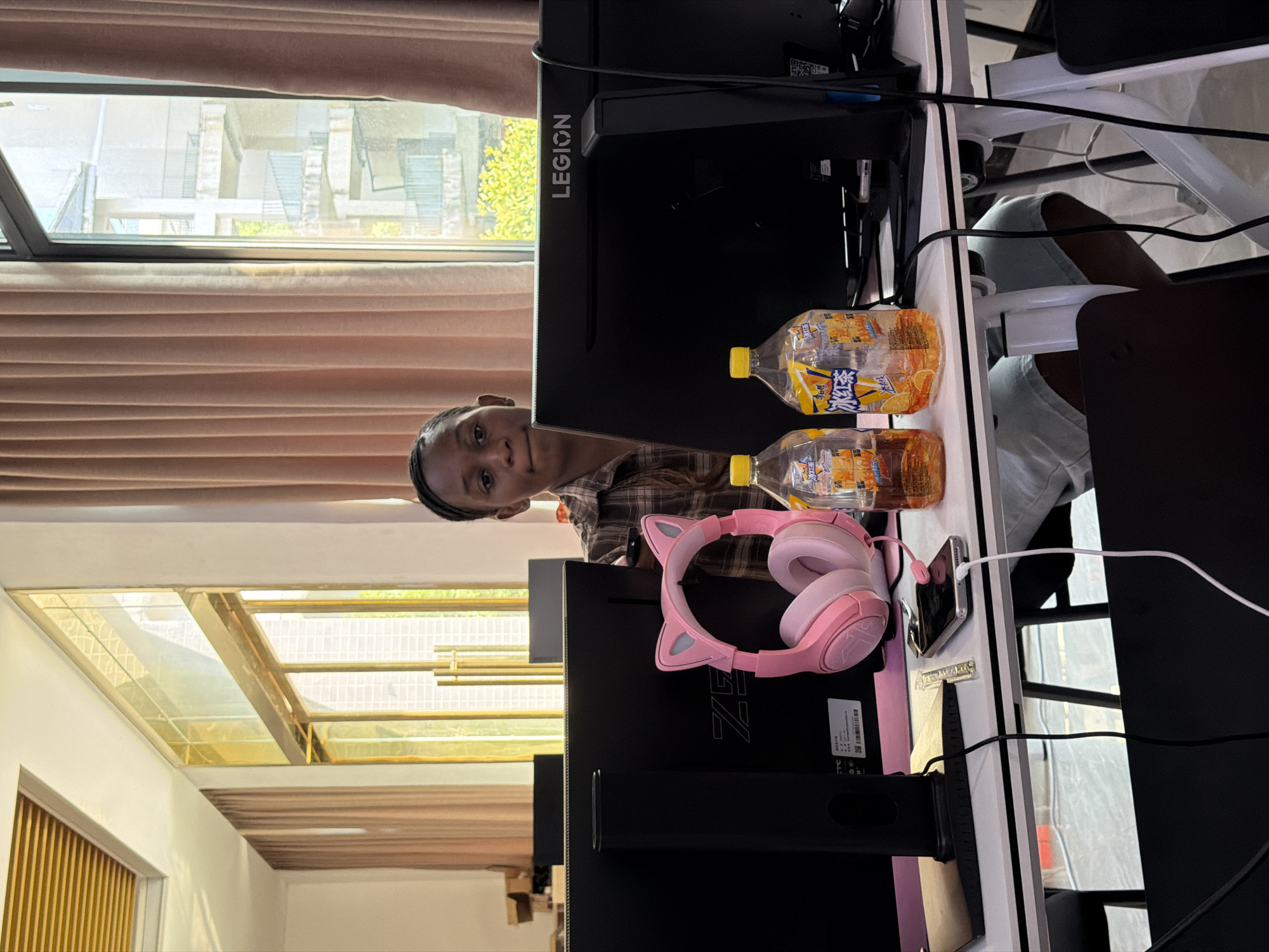A young woman sits at a desk with multiple computer monitors. She is wearing a plaid shirt and appears to be looking at the camera. On the desk are a pair of pink cat-ear headphones, two bottles of iced tea, and a smartphone charging. The background shows large windows with beige curtains, and an outdoor view of a building. The desk is white with black legs, and the surrounding environment suggests an office or computer lab setting. The overall impression is a casual work or gaming environment.