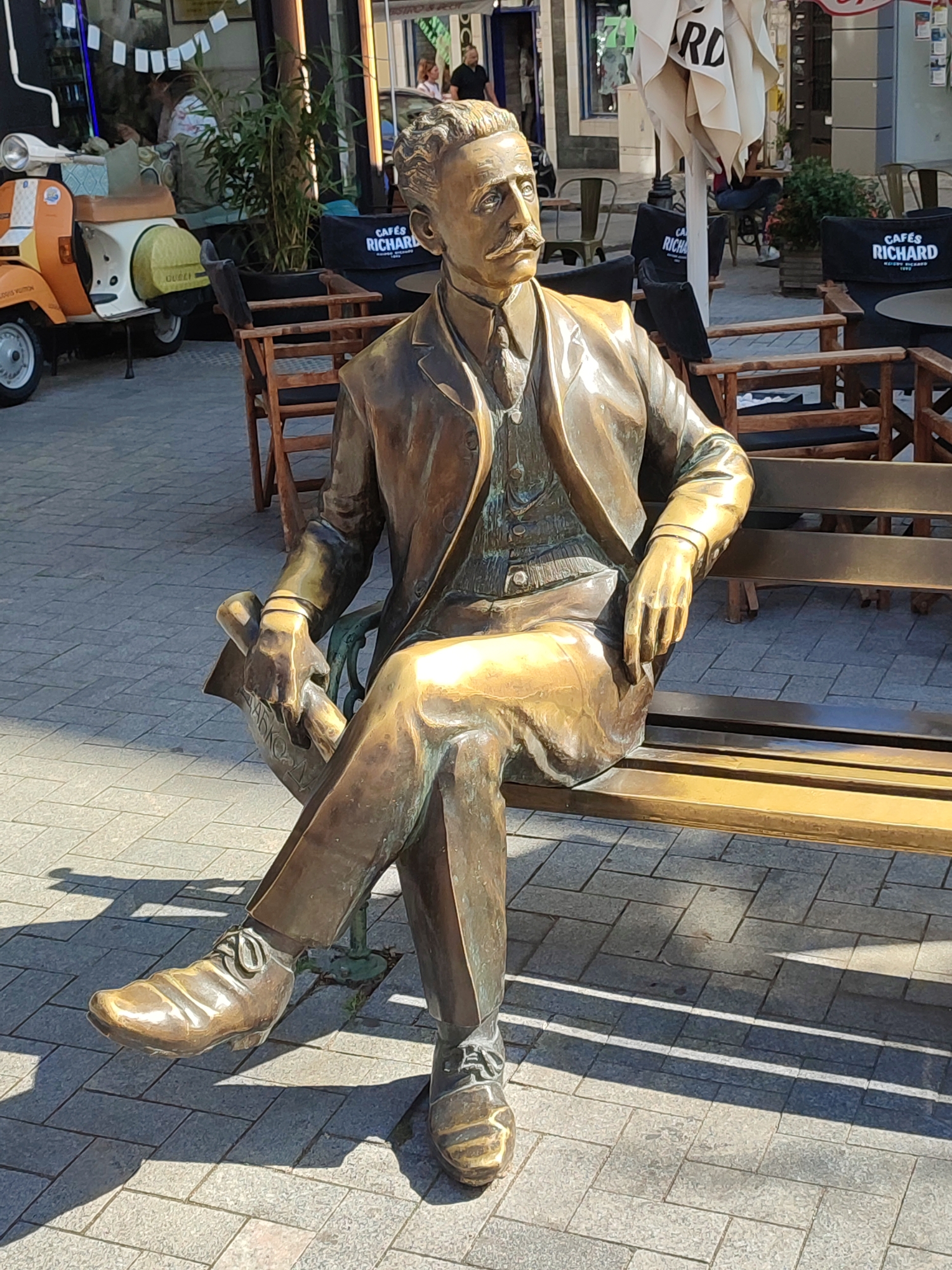 The image shows a bronze statue of a man sitting on a bench outdoors. The statue depicts a man in a suit, vest, and tie, with a mustache and curly hair. He is holding a rolled-up document or newspaper in his left hand. His right arm rests on his knee. The bench is made of wood and metal.
The statue is located on a paved pedestrian area. In the background, there is a scooter, outdoor seating with tables and chairs, and a building with an awning. Several black cushions with "CAFÉS RICHARD" written on them are visible on the chairs. The general ambiance suggests a cafe or public square setting.
Based on the presence of "CAFÉS RICHARD" outdoor seating and the style of the surrounding buildings and streetscape, the location is identifiable as the area near the entrance to the Sea Garden in Varna, Bulgaria, specifically at the intersection of Nezavisimost Square and Primorski Boulevard, near the monument to Georgi Dimitrov. The scooter appears to be a prop or part of the cafe's decor. The paving stones and the style of the cafe furniture are consistent with this area of Varna.