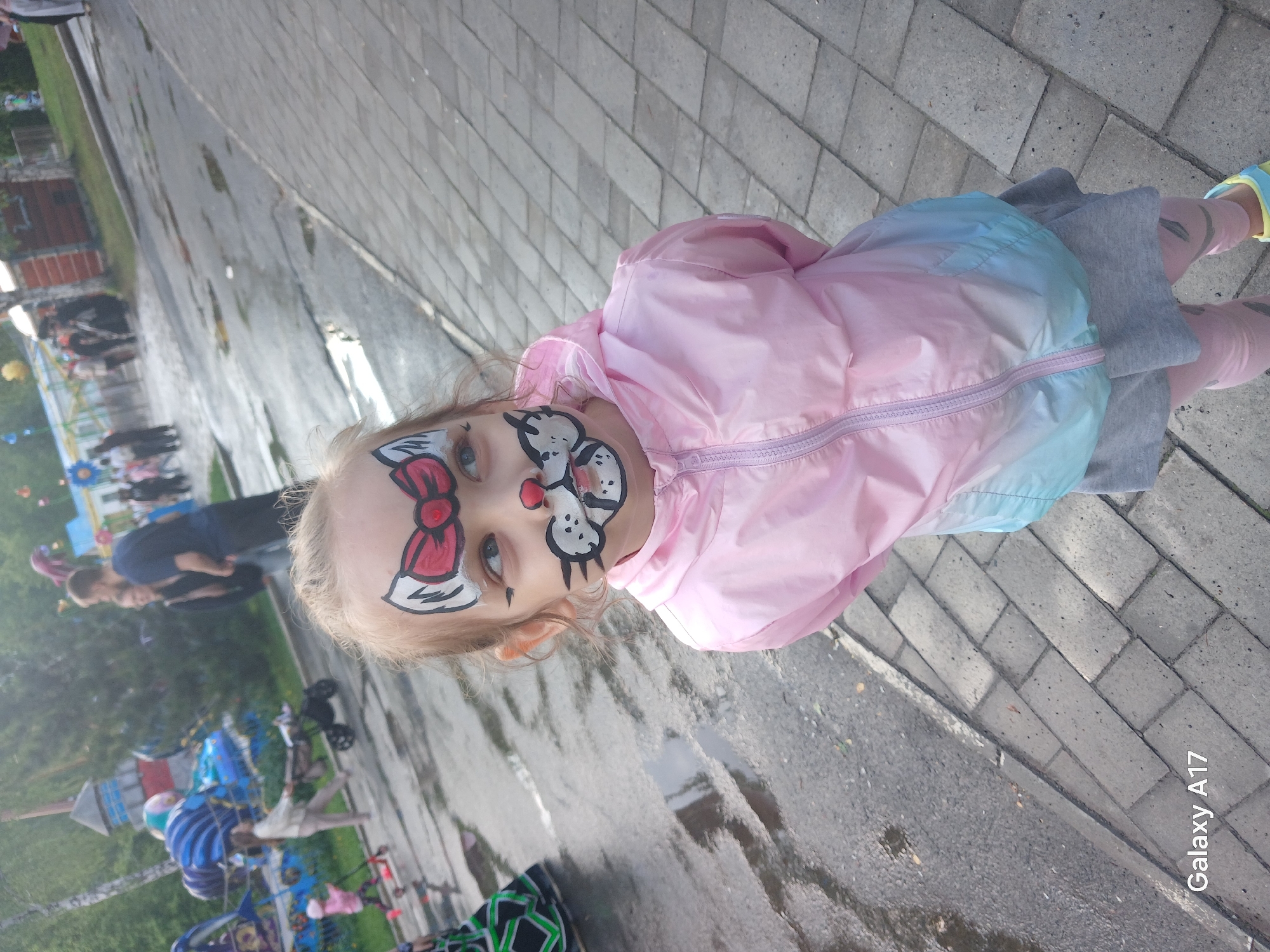 A young girl with face paint is standing on a paved area. She has a pink jacket with blue ombre on the lower half, and grey pants. The face paint includes a red bow on her forehead and cat whiskers and nose around her mouth. The background shows a park area with playground equipment and trees. People are present in the background, appearing to be other park attendees. The location is likely in the city of Yekaterinburg, Russia. The ground appears wet, suggesting recent rain. A "Galaxy A17" watermark is visible in the bottom right corner of the image.