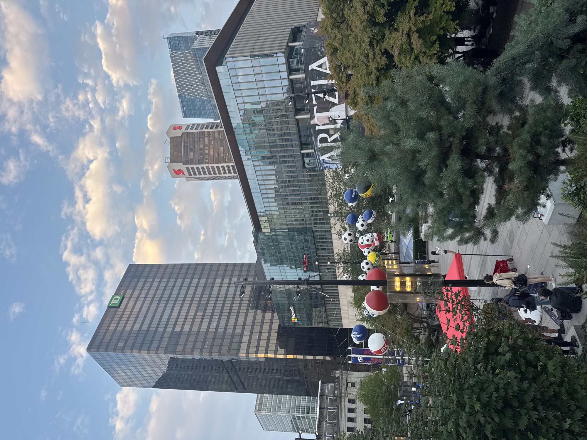 The image depicts a street scene in Vancouver, Canada, likely downtown, with modern skyscrapers dominating the skyline. In the foreground, a group of people are gathered near a red tent and a few trees. One person is seated on steps, while others stand and converse. Decorative spherical objects, resembling oversized beach balls and soccer balls, are suspended by wires above the pedestrian area, with some displaying country abbreviations like "USA" and "AUS".
The architecture features several prominent buildings, including a tall, dark glass skyscraper with a green TD logo at the top. Another building has a red stylized 'S' logo, likely for Scotiabank. A contemporary glass-fronted building displays a large banner for "ARITZIA". The sky is partly cloudy, with a soft, diffused light suggesting it might be late afternoon or early evening. The overall impression is a bustling urban environment with elements of public art or event decorations.
