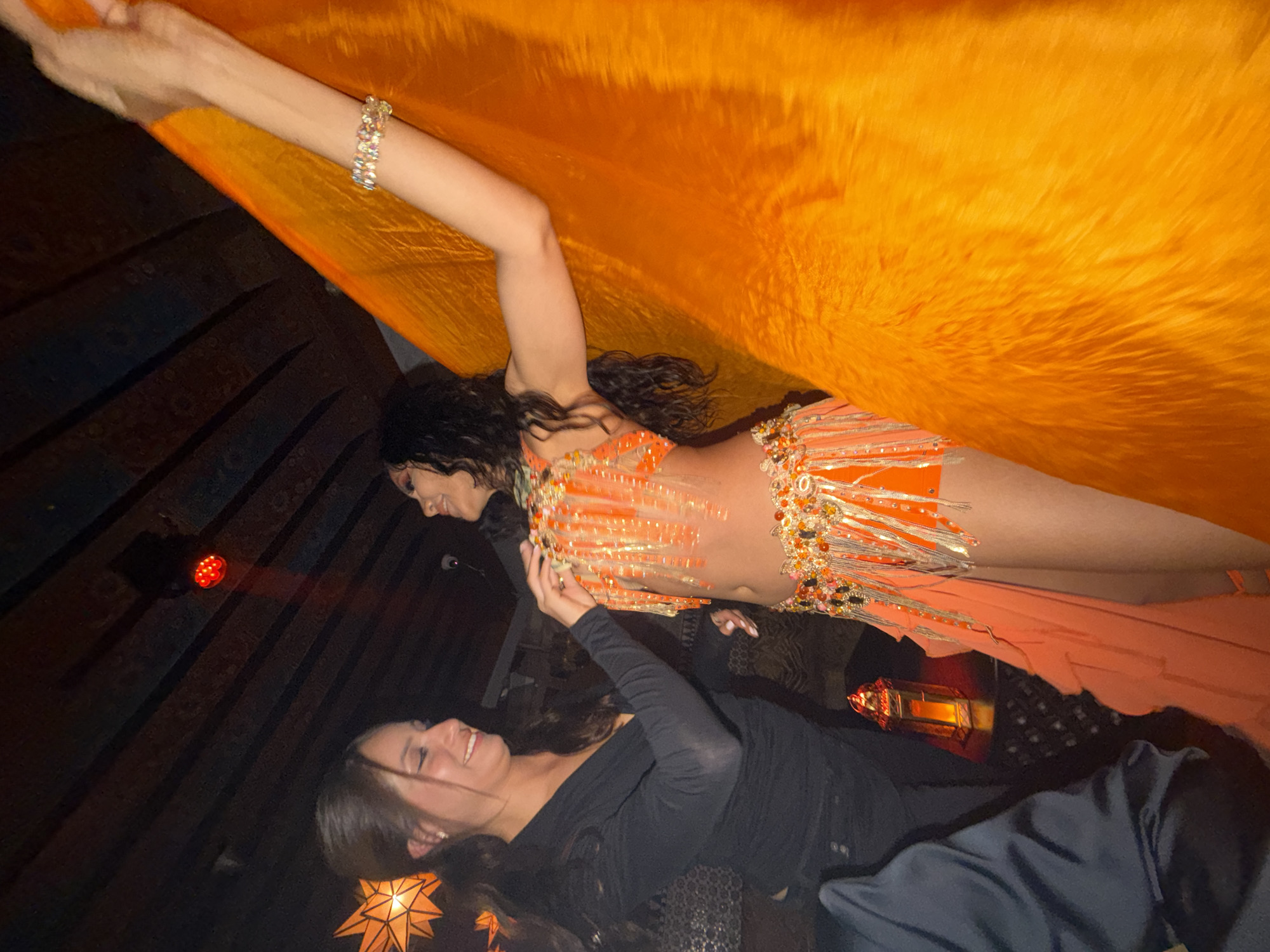 Here is a concise description of the image:
The image appears to capture a scene within an establishment. A woman in an embellished, orange belly dancing costume holds up a large orange veil with one arm, with a bracelet visible on her raised arm. A second woman is seen in the lower part of the frame, dressed in dark clothing, potentially interacting with the first woman or the veil. The setting includes dim lighting, with star-shaped light fixtures and a red stage light visible in the background. Given the presence of belly dancing and the overall aesthetic, this might be a themed party, performance, or event. There is no explicit information regarding the location.