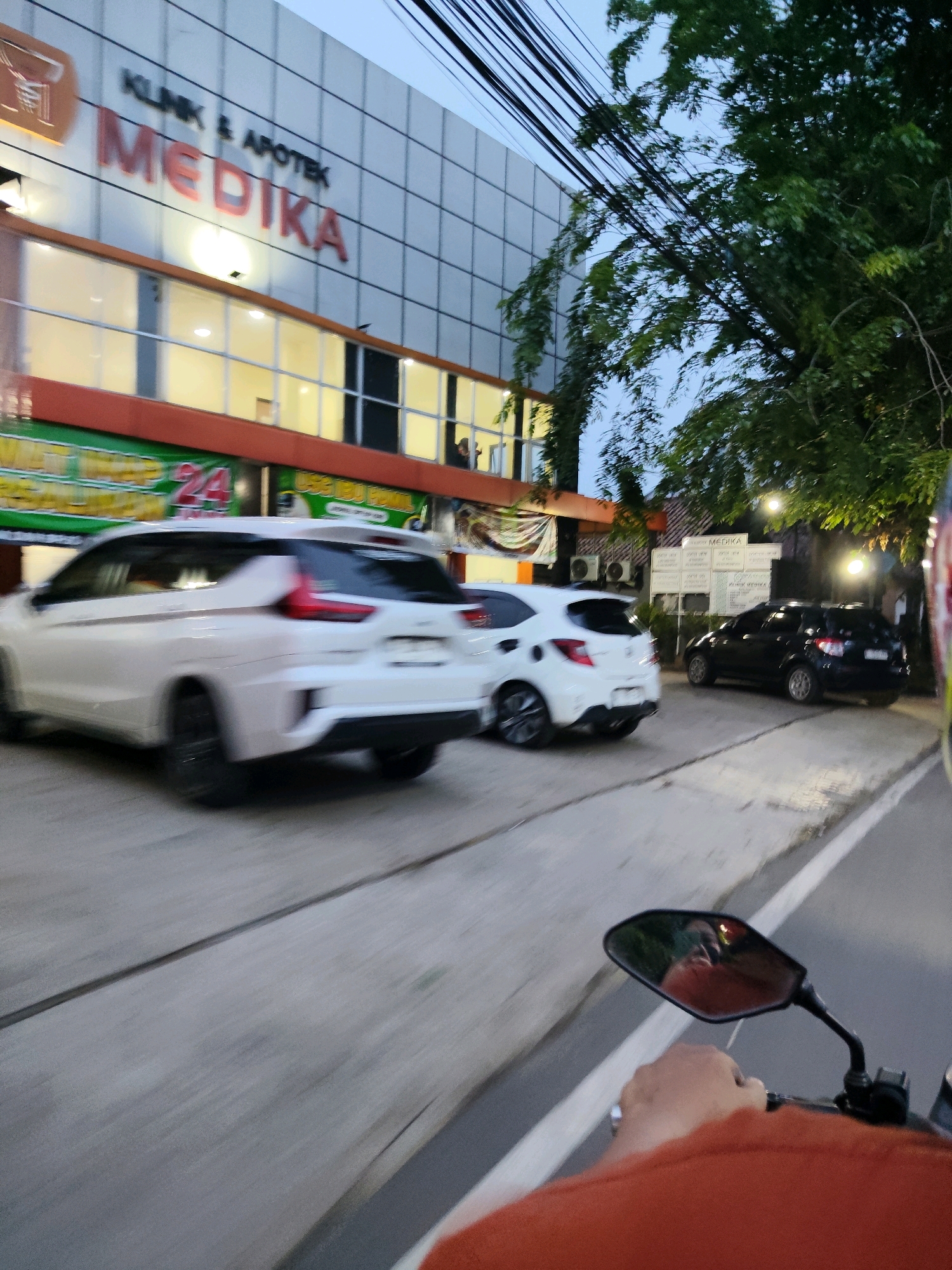 The image depicts a street scene in front of a building labeled "KLINIK & APOTEK MEDIKA" in Kaduagung Timur, Indonesia. A white car is in motion on the left side of the frame, and a black car is parked on the right. Trees and utility lines are visible overhead, suggesting an urban or semi-urban environment. The perspective indicates the photo was taken from a moving vehicle, likely a motorcycle, as a hand is visible on the handlebars and a mirror reflects the rider and the surroundings. The lighting suggests it is either dusk or early morning.
