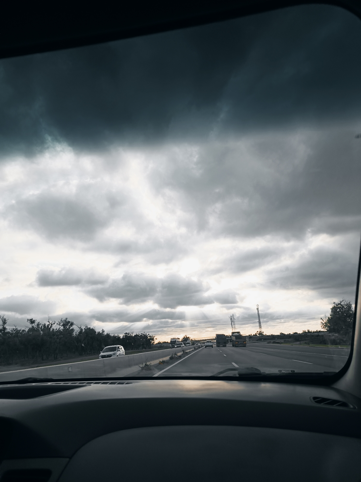 The image shows a view from inside a car driving on a highway. Outside, the sky is overcast with dark, heavy clouds. In the distance, there are electrical towers and vehicles ahead on the road. The landscape alongside the highway consists of low vegetation. The foreground is the dashboard of the car. The location is Shoul السهول, Morocco.