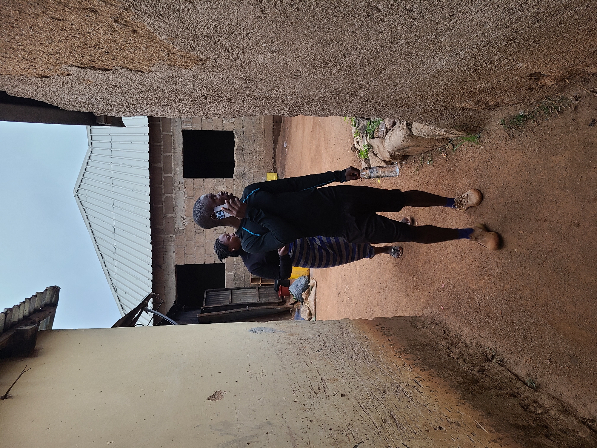 Two people are standing on a dirt path between two buildings. A man is on his phone and holding a plastic water bottle. A woman stands next to him. The environment is a compound in Jos, Nigeria.