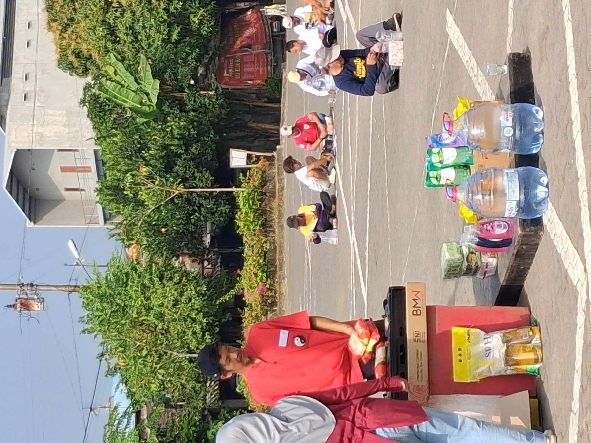 In the city of Surakarta, Indonesia, on an outdoor paved lot, a man in a red polo shirt with a yin and yang symbol is standing behind a black and red cart, holding a stack of yellow and red packaged goods. A woman in a gray hijab and a burgundy long-sleeved shirt stands to his left. In the background, several people are sitting on the ground in a semi-circle. Some are holding items and appear to be interacting with each other. Various items are displayed on the cart and on the ground around it, including bottles of water, packaged food items, and cleaning supplies. The environment suggests a casual gathering or a small market setup, with buildings and trees visible in the background under a clear sky.