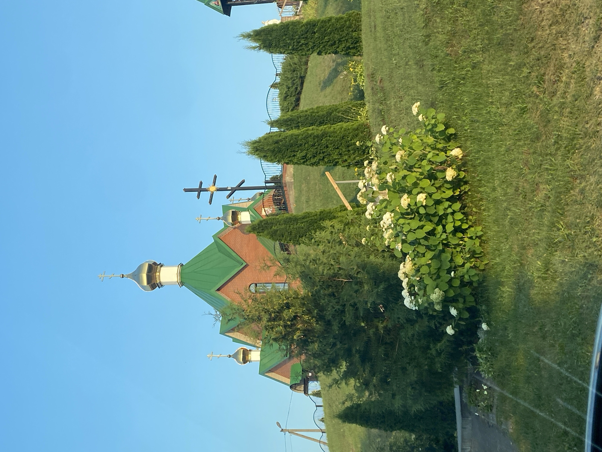 The image shows an Orthodox church with a green roof and golden domes, situated on a grassy hill. Several cypress trees and a blooming bush of white flowers are in the foreground. A cross is visible in front of the church. The sky is clear and blue. The scene is located in the village of Volchyn, which is part of the Volchynski Selsoviet in Belarus.