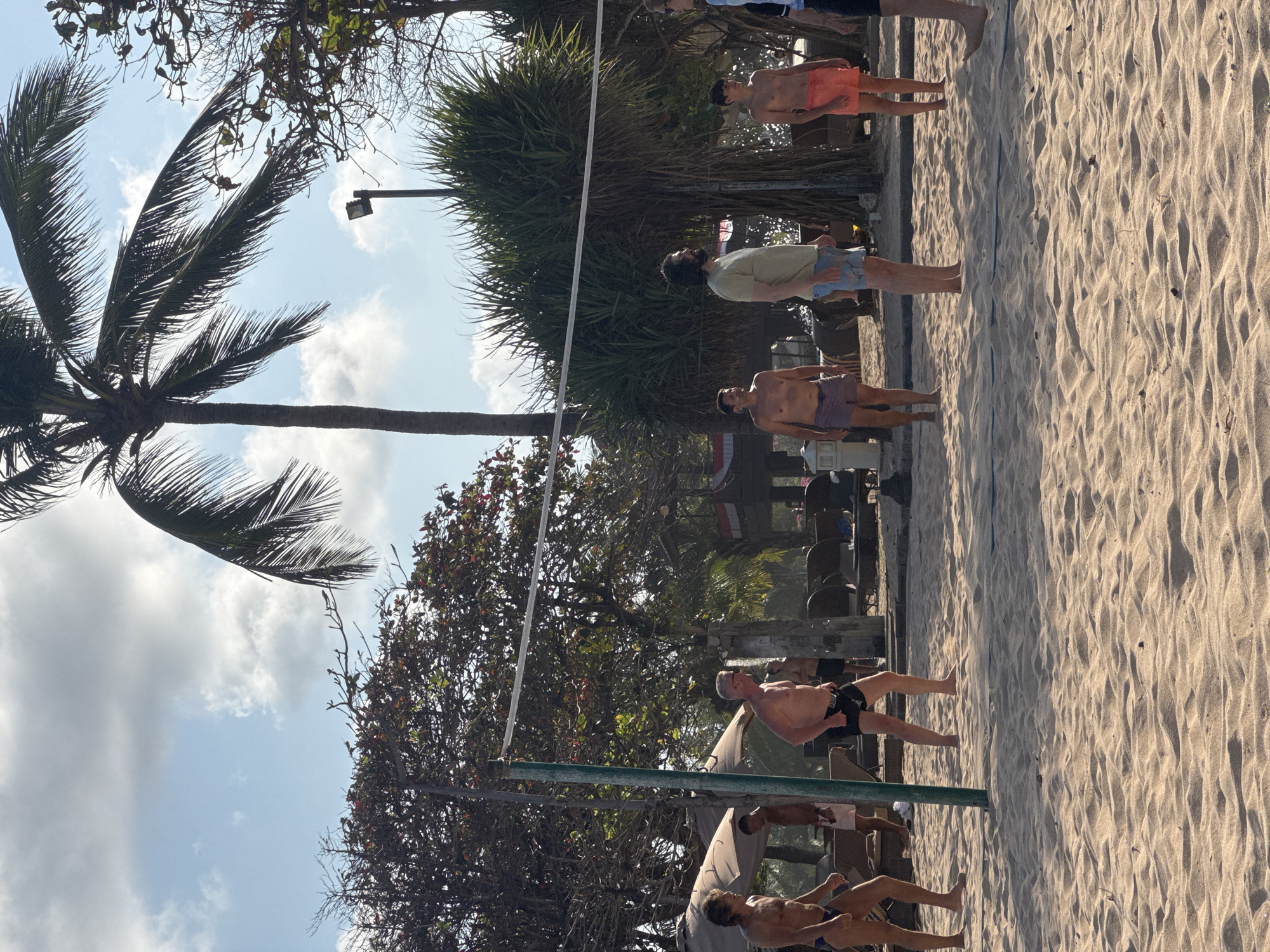 In Kuta Selatan, Indonesia, on a sandy beach, several men are playing volleyball. The setting is tropical, with palm trees and lush foliage visible in the background. The sky is partly cloudy, with a bright sun illuminating the scene. The men are casually dressed in swimwear, suggesting a relaxed atmosphere. One man on the left is wearing blue swim trunks, another in the center is in black shorts, and two others on the right are in orange and blue shorts respectively. The volleyball net is stretched across the court, with a green pole supporting one side. Beach chairs and umbrellas are scattered around, indicating a popular tourist destination. The sand is a light golden color, with visible footprints and undulations from the game.