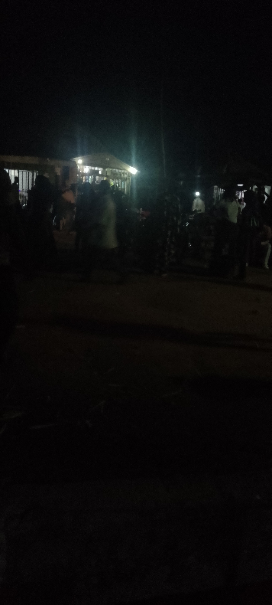 The image depicts a dimly lit outdoor scene at night, likely in Jalingo, Nigeria. In the background, a building with illuminated windows is visible, suggesting it might be a shop or a residential area. A group of people are gathered in front of and around the building. Their figures are mostly obscured by darkness and motion blur, making it difficult to discern specific details about their clothing or activities. Some individuals appear to be standing, while others might be walking or interacting. The ground in the foreground is dark and appears to be unpaved. The overall impression is of a lively, perhaps festive, gathering occurring after sunset.