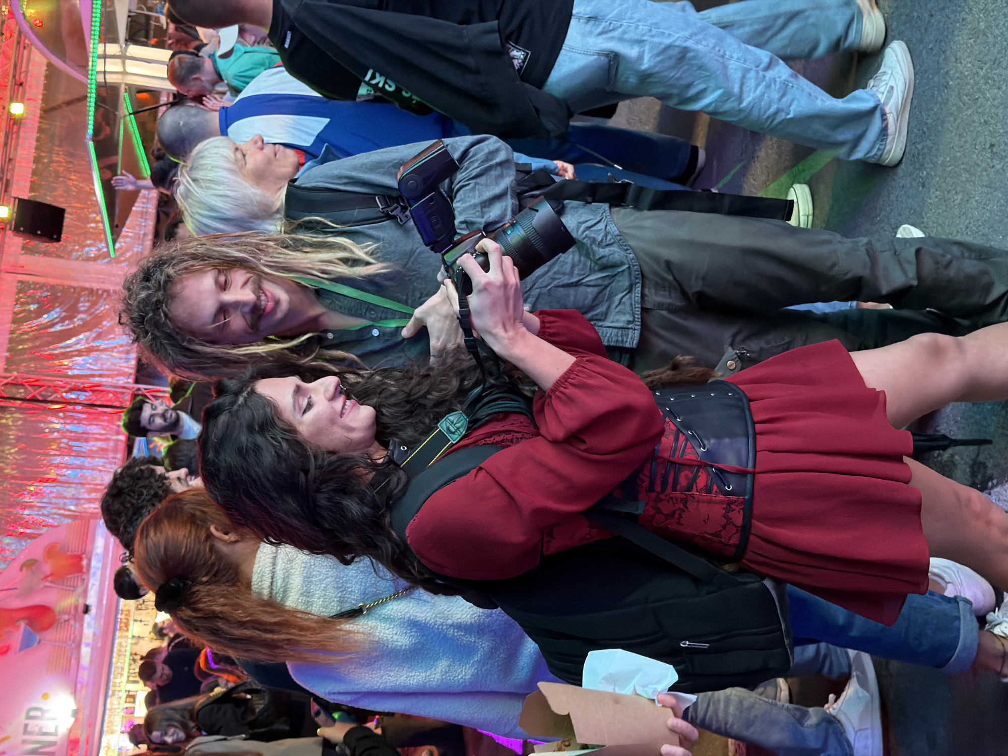 The image depicts a vibrant street scene at night, likely during a festival or outdoor event, possibly in Luxembourg City, Luxembourg, given the signage visible in the background which reads "LUX". In the foreground, a woman in a red dress and black corset is holding a camera, appearing to photograph a man with dreadlocks and a mustache. The man is wearing a grey button-down shirt and is also holding a camera. They are surrounded by a crowd of people, some of whom are blurred in the background. The environment is illuminated by colorful lights and decorations, suggesting a festive atmosphere. The people are dressed casually, in a variety of clothing styles. The overall impression is one of a lively social gathering, with photography being a prominent activity among the subjects.