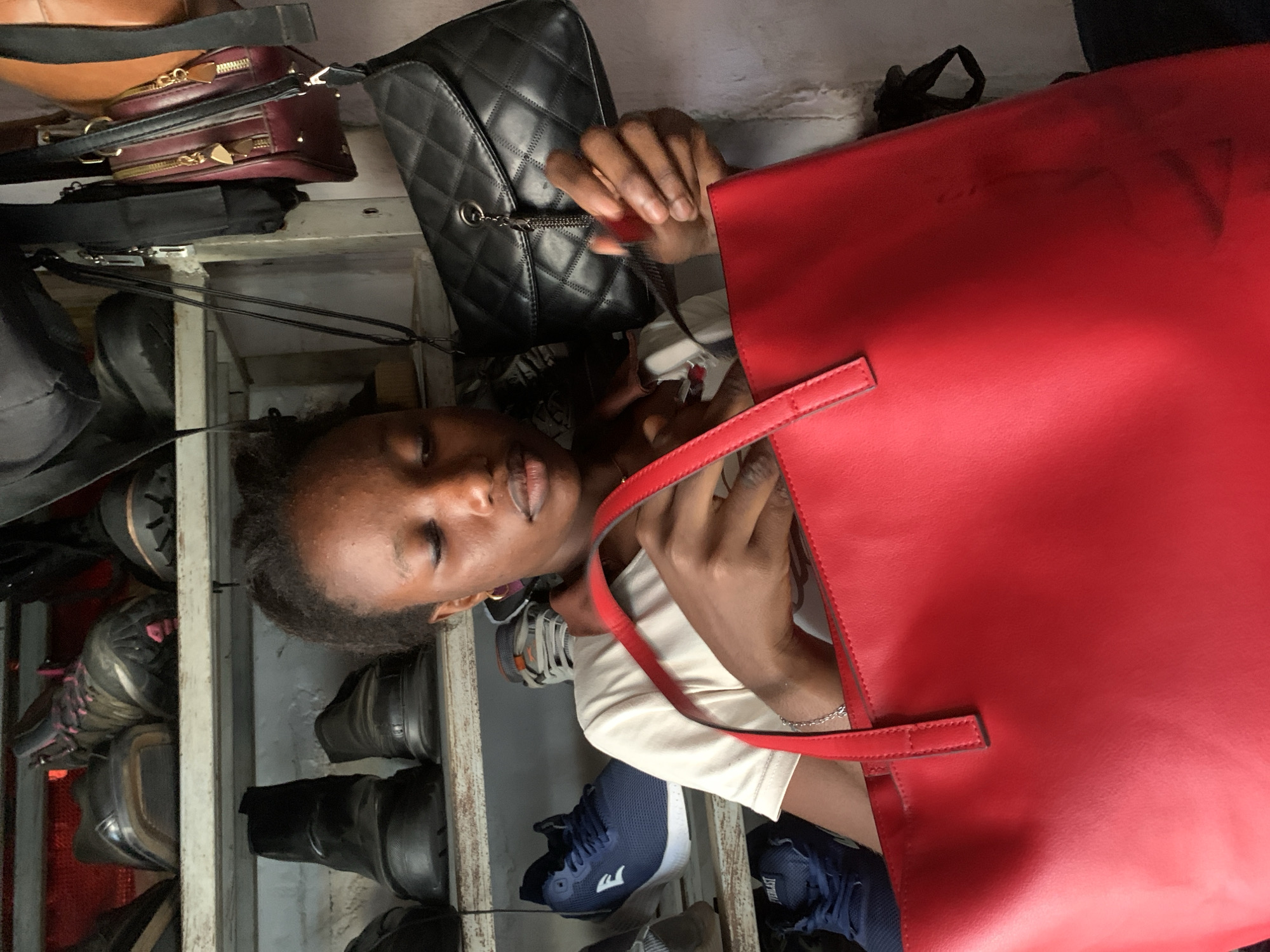 The image depicts a young Black woman in an indoor setting, likely a store or storage area, surrounded by shelves of footwear and handbags. The woman is in the foreground, holding a large red tote bag. She is wearing a light-colored t-shirt and has her eyes closed, concentrating on what she is doing with the bag. Behind her, shelves are stocked with various shoes, including boots and sneakers, and several handbags are visible, including a black quilted handbag and a burgundy bag with gold zippers. The environment appears somewhat cluttered, suggesting a retail or storage space. The lighting is natural, coming from an unseen source, and casts soft shadows. The image's composition focuses on the woman and the red bag she is holding. The location is identified as a store in Abuja, Nigeria.