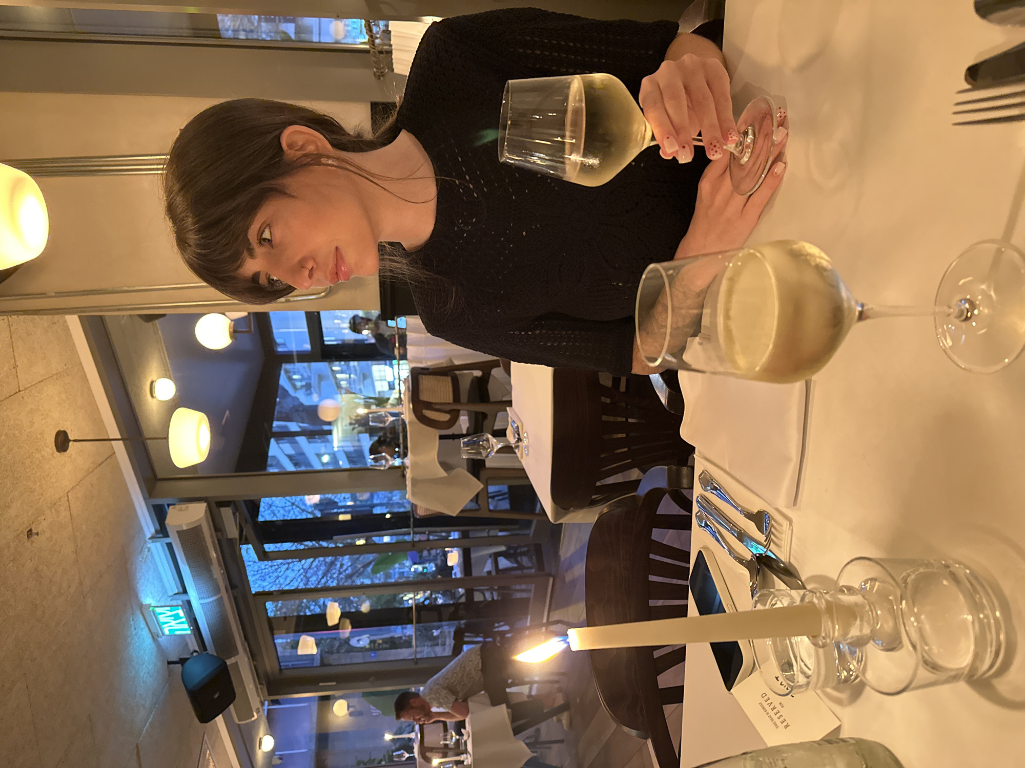 The image depicts an indoor scene in a restaurant or upscale dining establishment, likely during the evening in Tel-Aviv, Israel, given the dark views outside and the artificial lighting.
The primary subject is a woman seated at a table, holding a glass of white wine. She has dark hair with bangs and is wearing a black, patterned long-sleeved top. Her fingernails are adorned with a pink base and small dots. She looks towards her left with a subtle smile.
In front of her are two wine glasses, both containing a light-colored liquid, with one tilted on the white tablecloth. A lit candle in a clear glass holder casts a warm glow on the table, alongside cutlery and a small card that reads "RESERVED."
The background reveals a modern restaurant interior with large windows looking out onto an urban setting. The sky outside appears dark, indicating nightfall, with reflections of indoor lights and hints of distant buildings and possibly trees. Various light fixtures, including pendant lamps and wall sconces, illuminate the space. In the mid-ground, another man is visible leaning forward at a table. A green exit sign with Hebrew text, "יציאה" (Yetsiah), is mounted on a wall, reinforcing the Tel-Aviv location. An air conditioning unit is also visible near the windows. The overall ambiance is intimate and sophisticated, suggesting a quiet dinner or gathering.