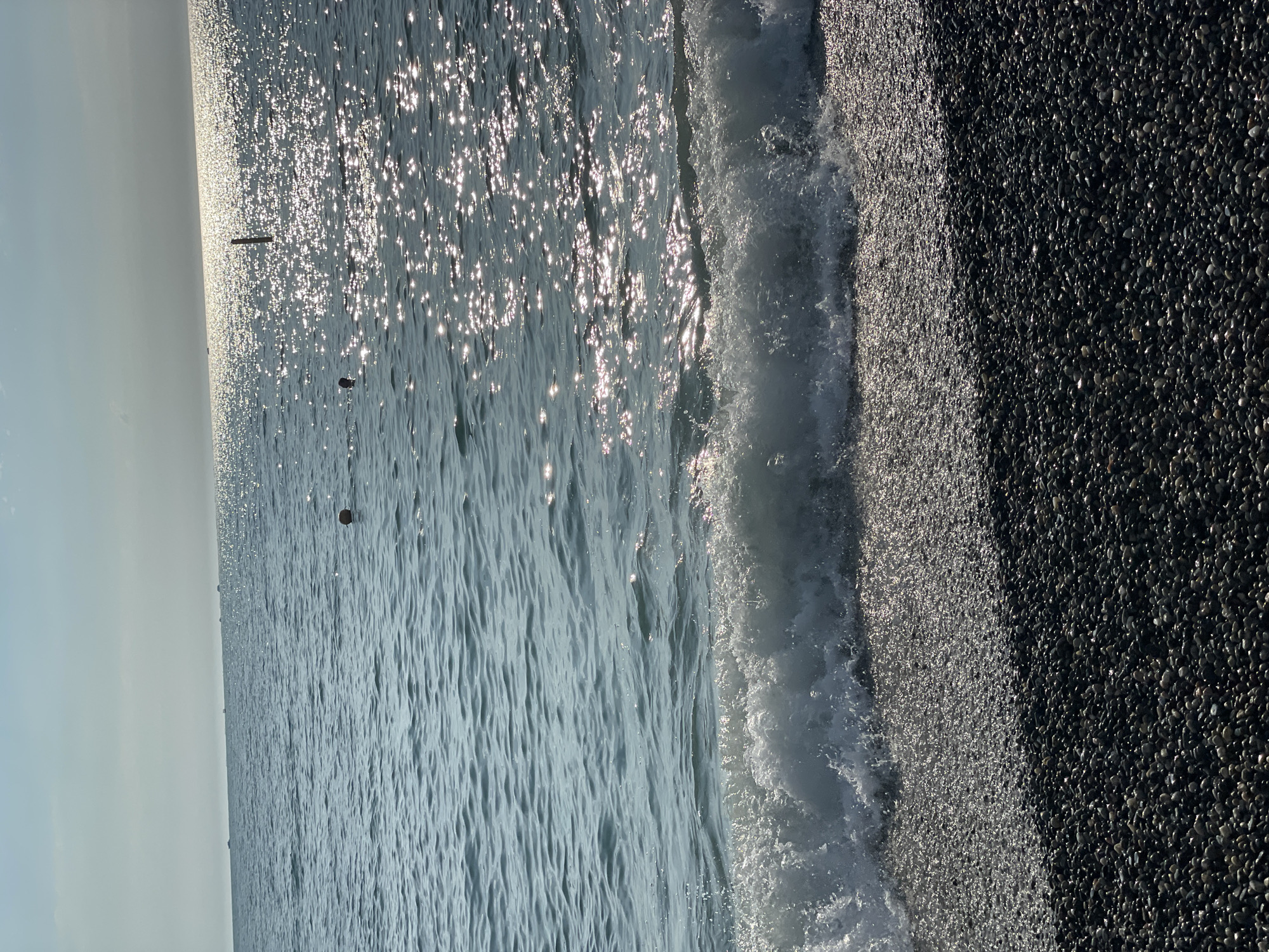 The image depicts a serene beach scene in Sochi, Russia. The sun's rays are glinting off the water, creating a dazzling effect on the surface. A gentle wave is breaking on the shore, which is covered in dark, smooth pebbles. The sky is a pale blue, and the overall atmosphere is peaceful and tranquil. The time of day appears to be late morning or early afternoon, judging by the angle of the sun and the brightness of the scene. There are no people visible in the image, and no text is present. The scene is natural and unadorned, focusing on the simple beauty of the coastline.