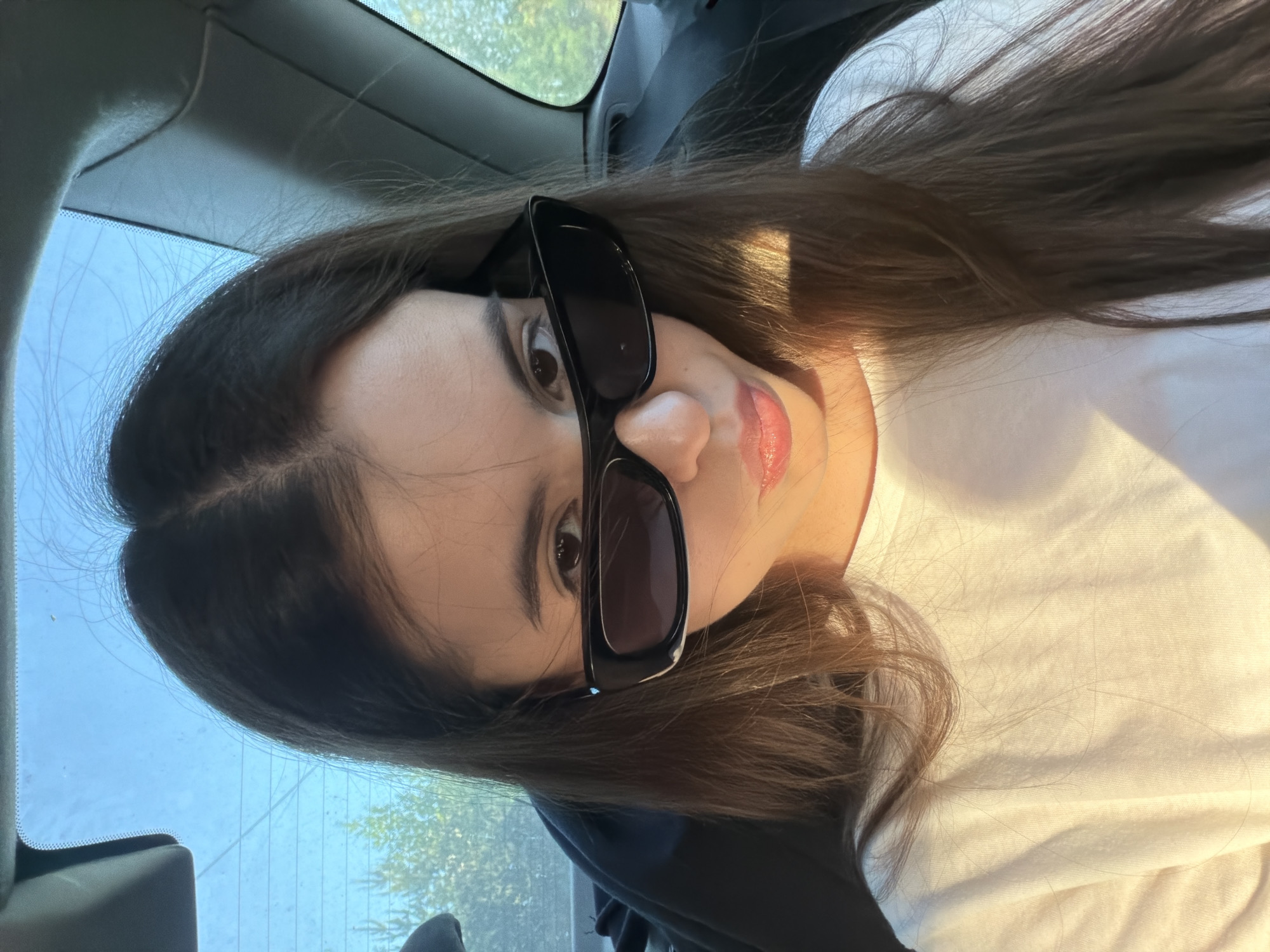 A young woman with long brown hair is sitting inside a car. She is wearing a white t-shirt and large black sunglasses. Light is streaming in from the right side of the image, creating a bright area on her shirt and face. The background shows a blurred view through the car window, including greenery and possibly other vehicles, suggesting an outdoor setting. The location within the city of Chaikovsky, Russia, cannot be determined from the provided image.