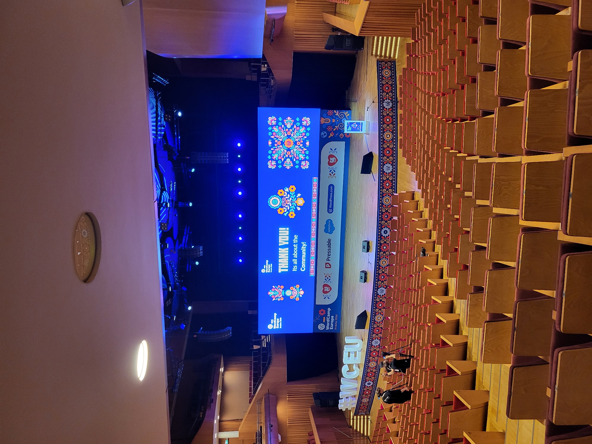 This vertical shot captures an expansive view of an auditorium or large conference hall, oriented with the stage on the left and rows of seating extending to the right. The hall is mostly empty of attendees, suggesting it's either before or after a main event, or during a rehearsal or setup.
The prominent feature is a large, brightly lit LED screen on the stage, displaying a blue background with colorful, folk art-inspired floral patterns. The screen features the text: "THANK YOU! It's all about the Community!" and multiple instances of the "WordCamp Europe" logo. Below the main thank-you message, additional logos for sponsors or partners are visible, including "Pressable," "Salesforce," and "WordPress.com." A smaller screen or podium near the center of the stage further announces "WordCamp Europe Kraków 2026," indicating a future event.
The seating area comprises numerous rows of auditorium-style chairs with wooden backs and what appear to be red or maroon upholstered seats. A long, elaborately patterned rug or runner, featuring traditional floral and geometric designs in red, blue, and yellow hues, stretches along the aisle towards the stage.
On the stage floor, equipment such as speakers and stage lights are visible. In the lower right corner of the frame, near the edge of the stage and the seating, a few individuals, likely event staff or crew, are present, some appearing to operate cameras or other technical equipment. The hashtag "#WCEU" is also visible on the stage floor in large letters. The overall lighting is a mix of bright blue and white stage lights contrasting with softer ambient lighting in the seating area.