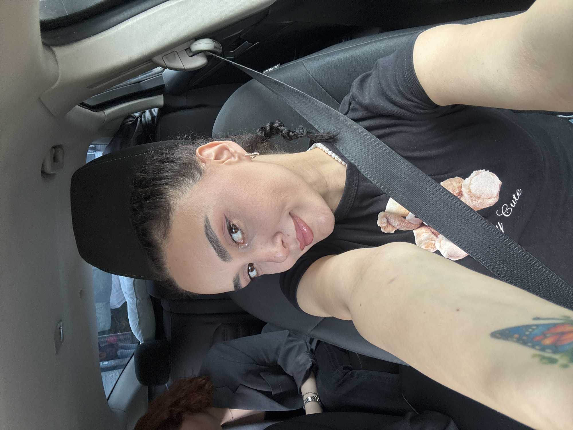 A female with fair skin, brown hair with braided sections, wearing a black t-shirt with a teddy bear graphic and the word "Cute," is seated in a car and wearing a seatbelt. The interior of the car features black leather seats and gray ceiling panels. The woman has a butterfly tattoo on her right forearm and is wearing a pearl necklace. Her face is turned towards the camera. Another person with reddish-brown hair is partially visible in the lower left corner. Outside the car window, items including clothing are visible. The location is Tbilisi, Georgia.