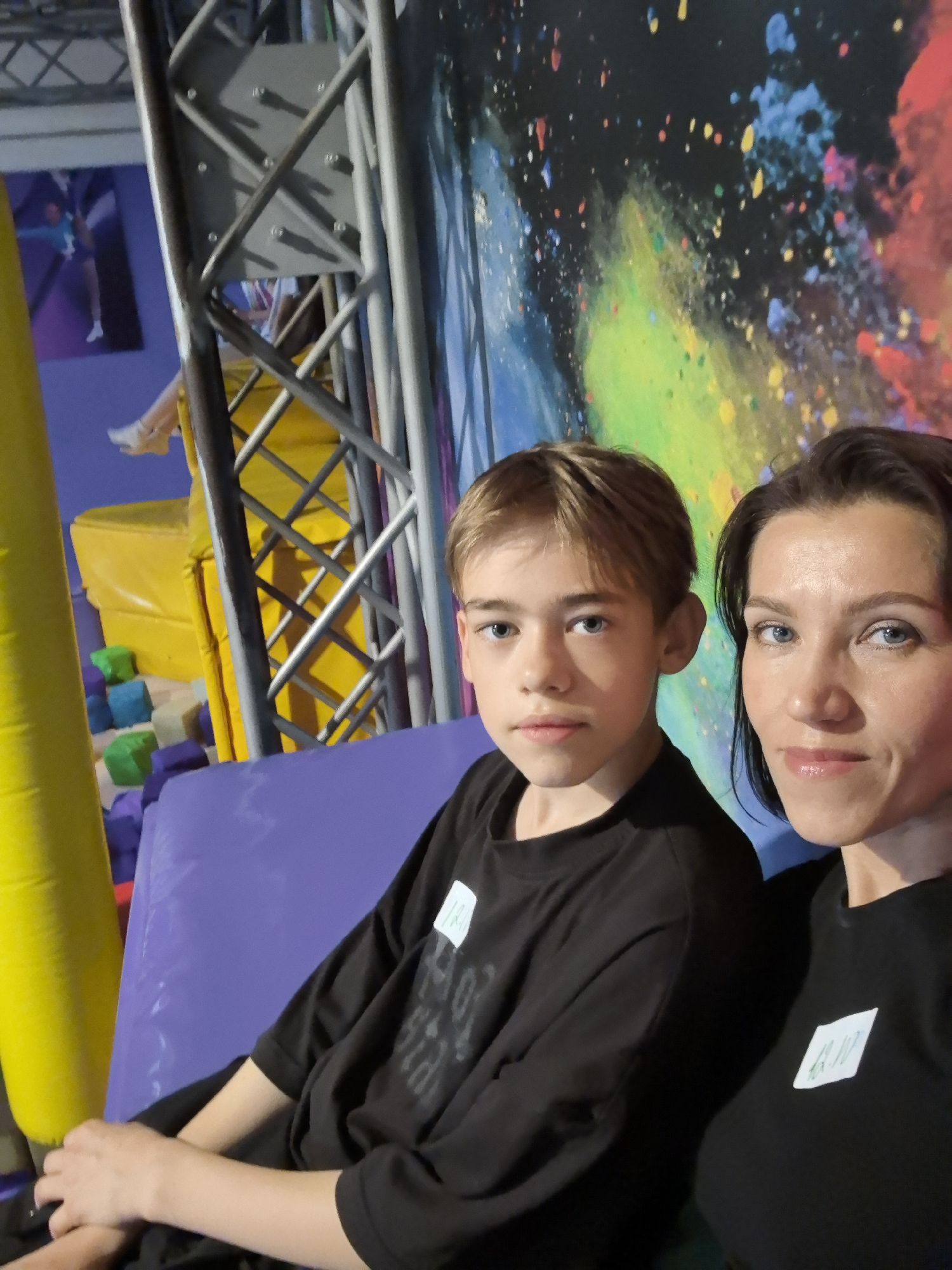 A medium shot captures a boy and a woman posing for a selfie in what appears to be an indoor entertainment venue. The boy, positioned on the left, has light brown hair and blue eyes, wearing a black t-shirt with a white tag. The woman, on the right, has dark brown hair and blue eyes, also wearing a black t-shirt with a white tag. Both are looking at the camera with neutral expressions. The background features a colorful mural resembling exploding paint or powder, with abstract shapes in red, yellow, green, and blue against a dark background. To the left, a large yellow inflatable object and part of a metal structure are visible, along with a purple mat. The overall scene suggests a recreational or activity-based location, possibly an indoor trampoline park or similar establishment. The precise location in the city of Perm, Russia, cannot be determined from the visual information alone.