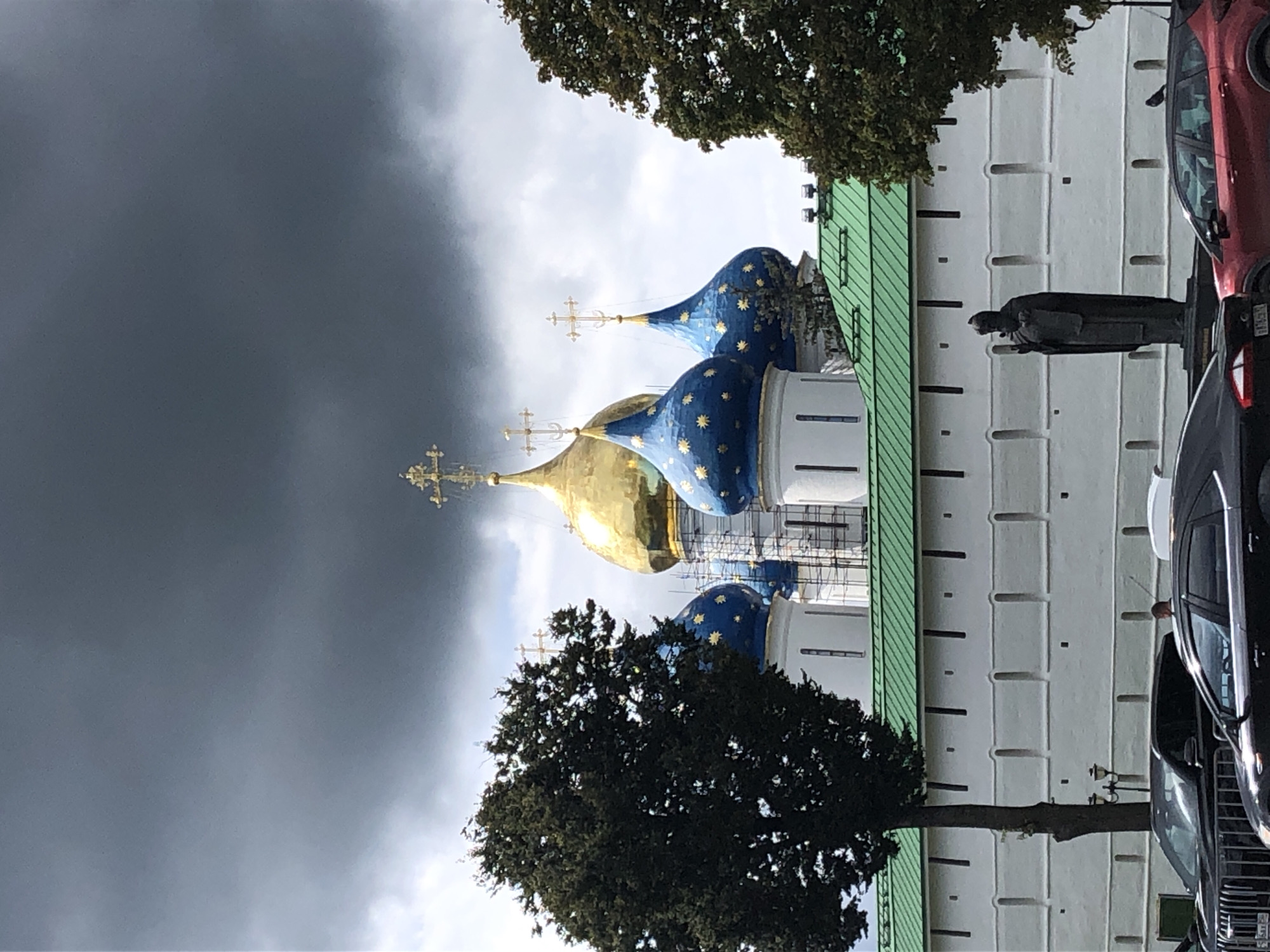 The image depicts the Trinity Lavra of St. Sergius in Sergiev Posad, Russia, under a dramatic, overcast sky. The primary subjects are the distinctively shaped onion domes of the lavra's cathedrals, one large golden dome crowned with a cross, and several smaller blue domes adorned with gold stars, also topped with crosses. These structures are partially obscured by scaffolding, suggesting ongoing renovation or maintenance. In the foreground, a white wall with decorative vertical indentations runs horizontally across the frame, topped by a bright green roof. To the left of the wall stands a tall, dark green tree. On the right, another dark green tree frames the scene. A statue of a robed figure, likely a saint, stands in front of the white wall. Several cars are visible in the lower part of the image, parked along a street in front of the lavra's wall. A single bird is perched on the roof of a red car. The overall impression is one of historical grandeur contrasted with the imposing, moody sky and the mundane presence of modern vehicles.