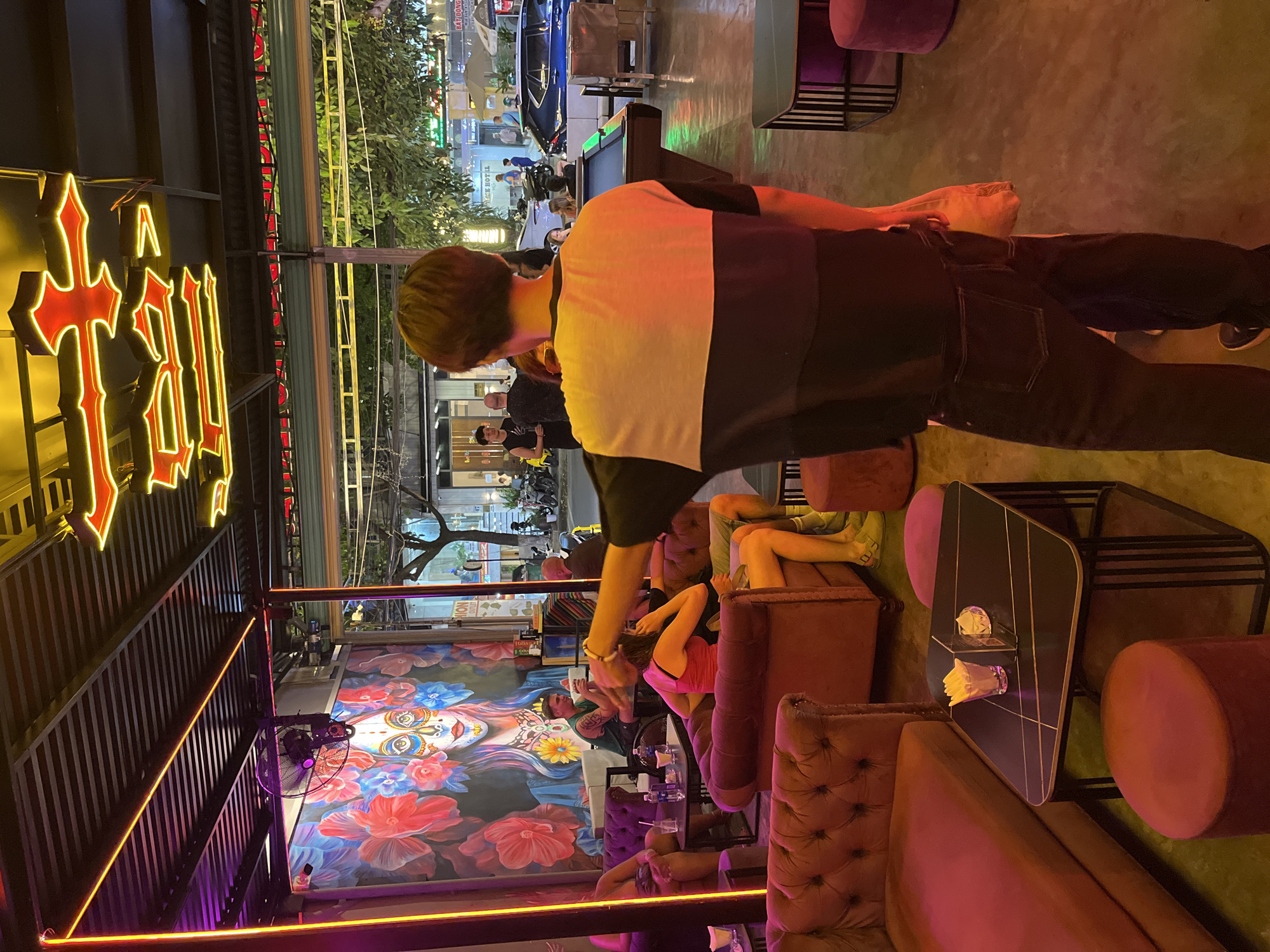 The image depicts a nighttime scene at a bar in Đà Nẵng, Vietnam. Patrons are seated on velvet sofas, illuminated by neon lights and enjoying the ambiance. A pool table is visible in the background. The bar features vibrant floral murals on the walls. The image is taken from an elevated perspective, with a man in a striped shirt partially in the foreground.