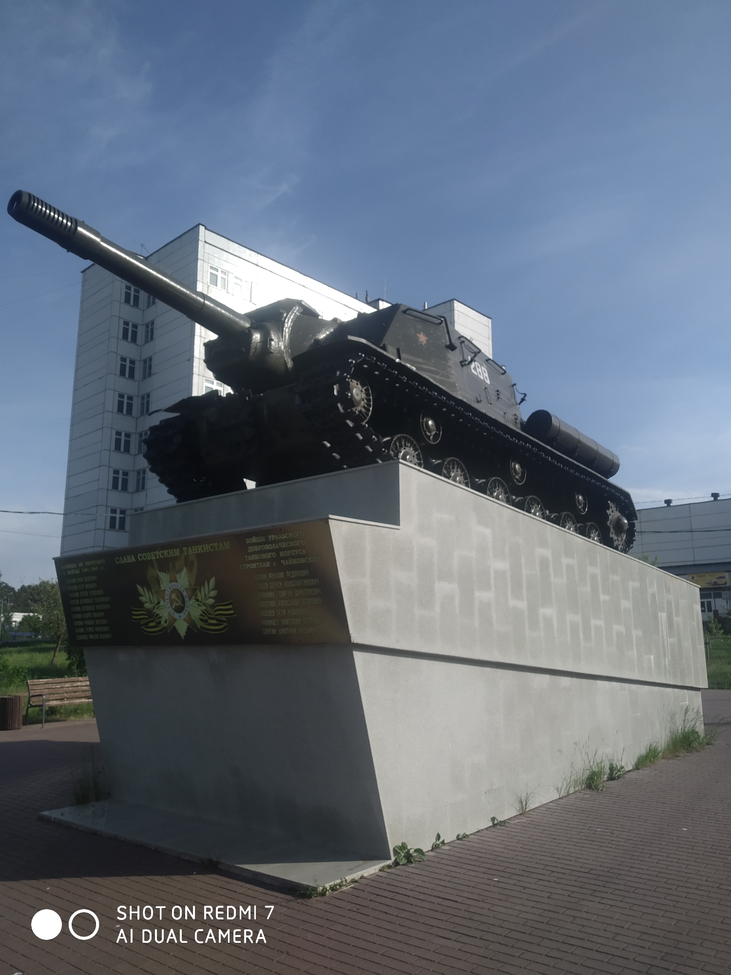 A military tank mounted on a concrete pedestal is the focal point of the scene. The tank is painted in a dark color, with the number "288" visible on its side. A building rises in the background. The pedestal has a bronze plaque on one side that reads in Russian: "СЛАВА СОВЕТСКИМ ТАНКИСТАМ" (Glory to Soviet Tankers) along with other text in Russian and features a Soviet-era emblem in the center. The pedestal is located in the city of Chaikovsky, Russia, which is inscribed on the plaque in Russian as "СТРОИТЕЛИ. ЧАЙКОВСКИЯ" (Builders. Tchaikovsky). The tank is positioned on a paved walkway. There is a lawn with benches and trees to the left of the monument. The weather is sunny. The photograph was taken with a Redmi 7 phone, indicated by the watermark at the bottom.