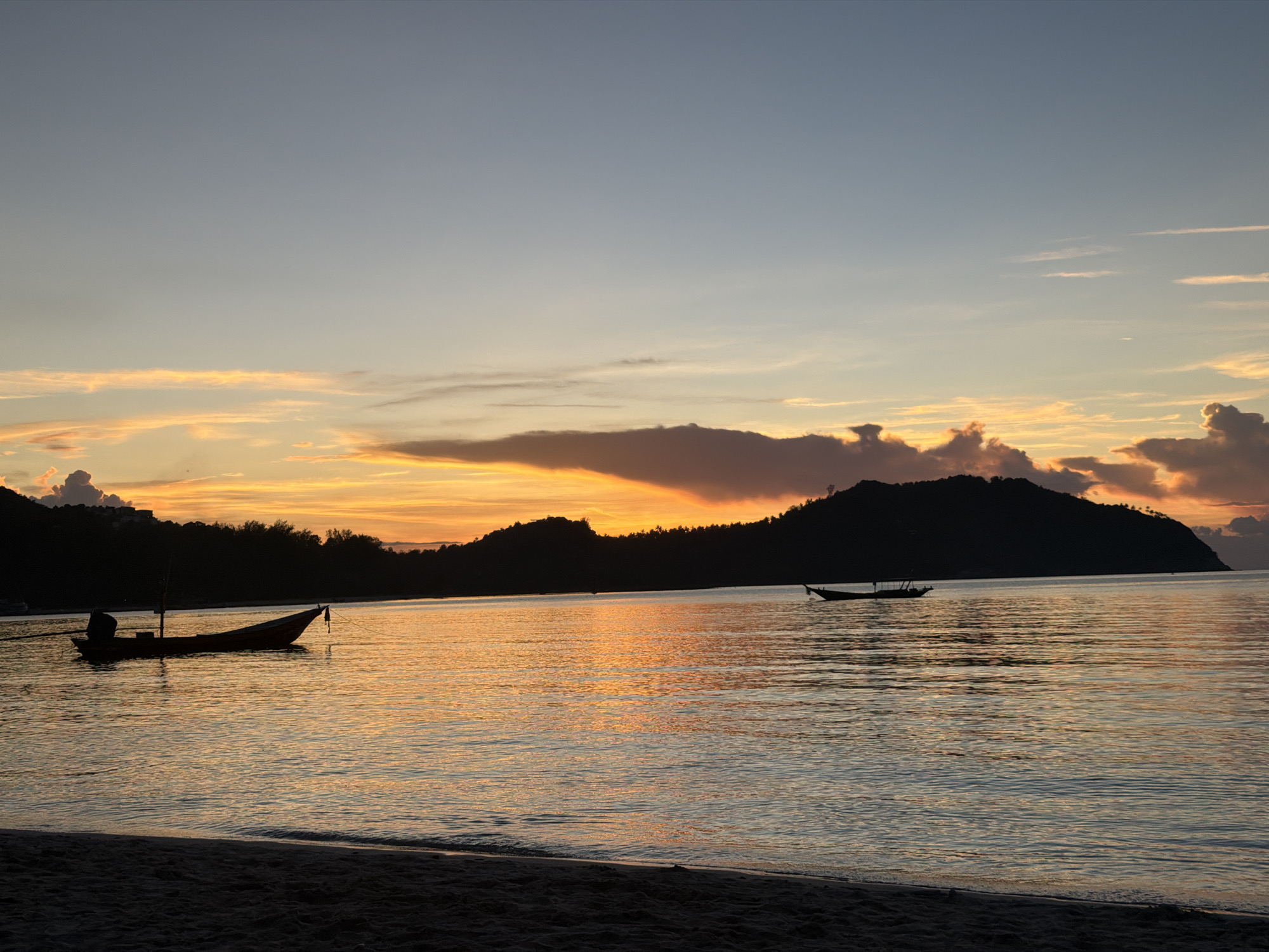 The image depicts a serene sunset scene on the coast of Koh Phangan, Thailand, specifically within the district of Tambon Ko Pha Ngan. The sky is a gradient of soft blues and oranges, with streaks of vibrant yellow and pink from the setting sun illuminating the clouds. The calm sea reflects these warm hues, creating a shimmering, golden surface.
In the foreground, a sandy beach is visible, dark and silhouetted against the brighter water. Two traditional longtail boats are present in the water. The closer boat, on the left side of the frame, is moored and appears empty, with a dark silhouette against the water. Further out in the distance, another, smaller boat is visible as a dark shape on the horizon.
Dominating the background are the dark silhouettes of rolling hills or islands covered in dense vegetation. The outline of these landforms is sharp against the colorful sky. The overall impression is one of tranquility and natural beauty, characteristic of a tropical sunset.