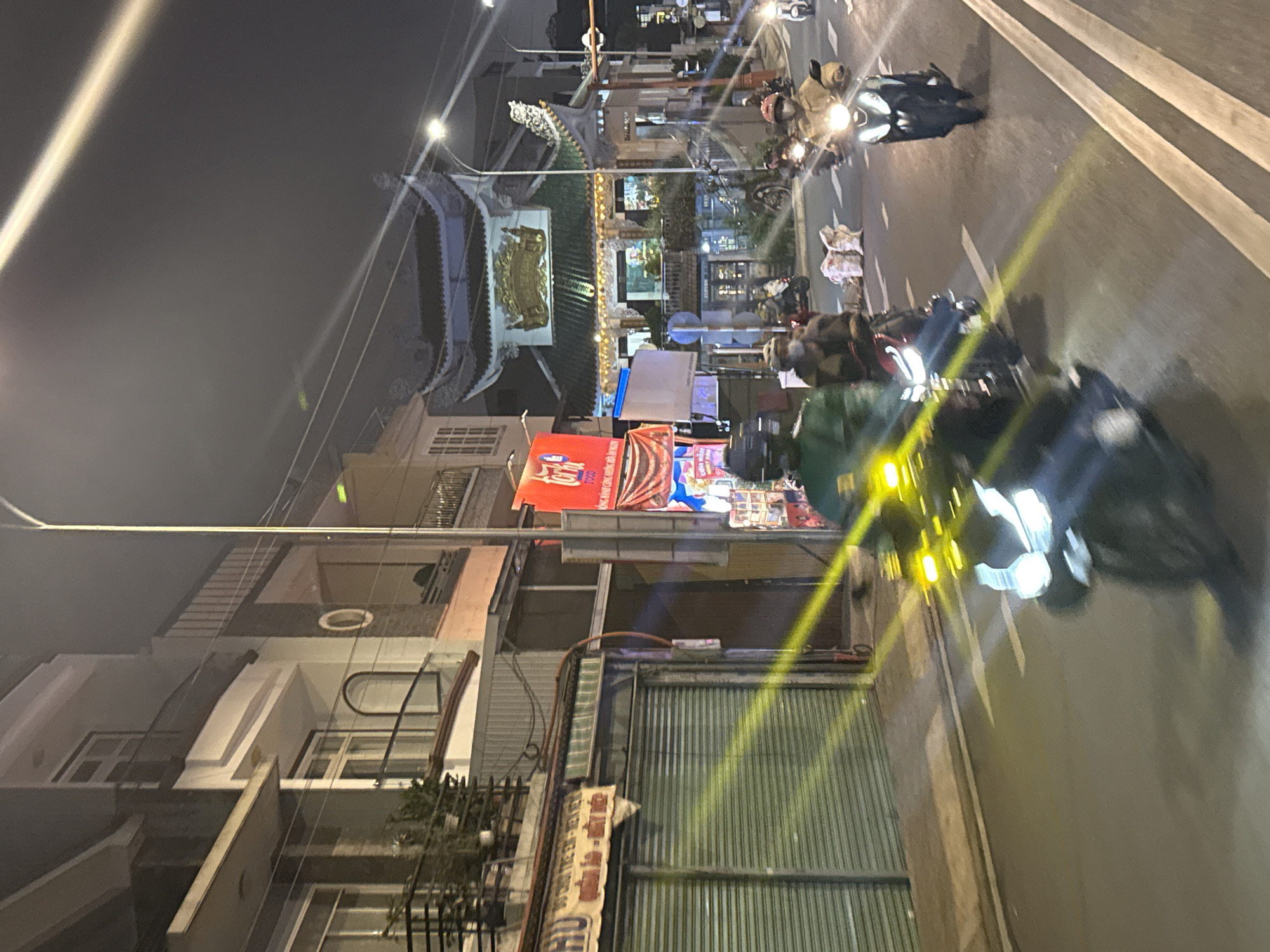 This media file captures a vibrant night scene on a street in Cam Ly, Đà Lạt, Vietnam. The street is bustling with activity, primarily featuring numerous motorbikes, many with their headlights brightly illuminated, creating streaks of light and glare across the asphalt road. Several individuals can be seen riding or standing near their motorbikes.
The street is lined with multi-story buildings. A prominent architectural feature on the right side of the street is a traditional Vietnamese-style gate or temple entrance, characterized by its ornate green tiled roof and a decorative golden plaque or sign above the main entryway. Other buildings include modern structures and those with corrugated metal rolling doors. Overhead power lines are visible against the dark night sky.
Artificial lighting is pervasive, coming from streetlights, vehicle headlights, and illuminated commercial signs. On the left side of the street, a large red sign clearly displays the English word "FOOD," accompanied by Vietnamese text below it, likely advertising food or snack items. Another banner with Vietnamese text is partially visible further down.
The time of day is clearly night, with a dark sky and the entire scene dependent on artificial light sources. No specific weather conditions are discernible, suggesting a clear night. The image portrays a typical lively urban street scene at night in Vietnam, characterized by heavy motorbike traffic and commercial activity.