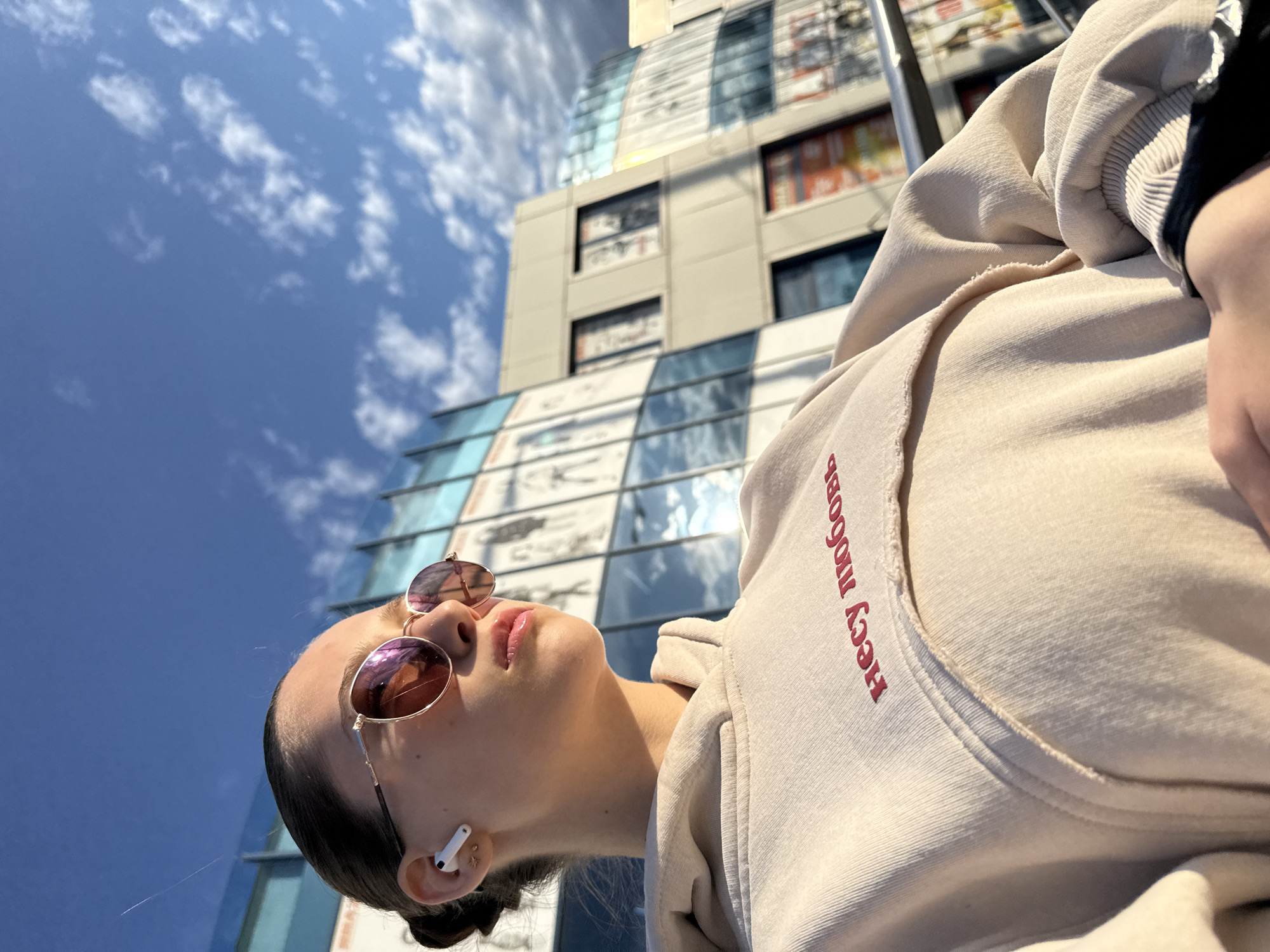 A low-angle, wide shot captures a young woman with sunglasses and an AirPod in her left ear. She is wearing a beige hoodie with red Cyrillic lettering. Her head is tilted back, facing towards the sky. Behind her is a modern building with a blue and grey façade, partially obscured by the bright blue sky with scattered clouds. The location is the city of Sofia, Bulgaria.
