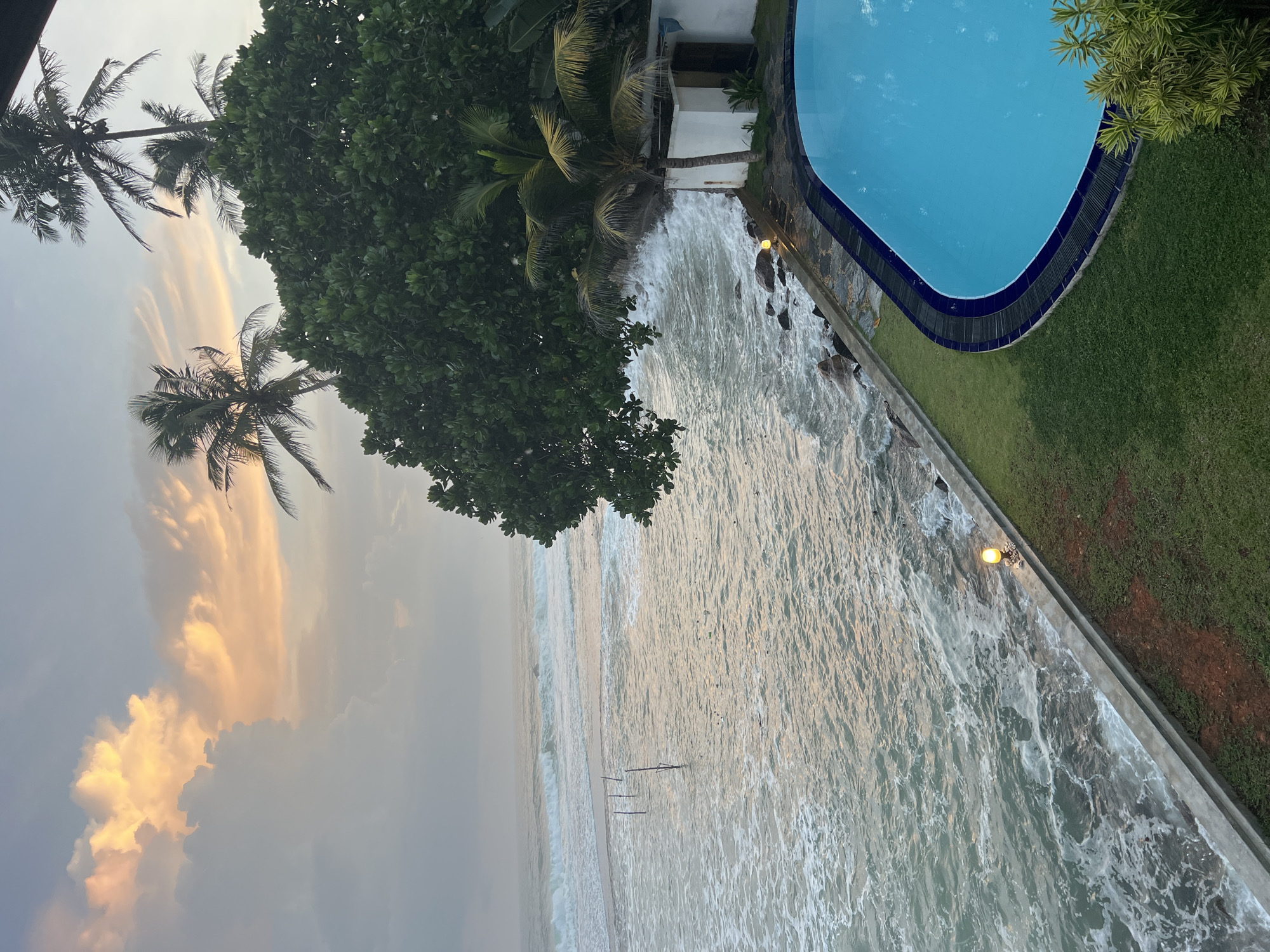 The image depicts a coastal scene at dusk in Goviyapana, Sri Lanka. A bright blue swimming pool occupies the foreground on the right, bordered by lush green grass. Adjacent to the pool is a white structure with a brown door and a small plant. Waves crash against the rocky shoreline to the left of the pool. Further left, the ocean extends to the horizon, with waves and foam visible. Several thin wooden poles are planted in the water, a common sight for stilt fishing. The sky above is filled with dramatic clouds, tinged with orange and yellow hues from the setting sun. Tall palm trees and dense green foliage are scattered throughout the scene, framing the ocean and sky. The overall atmosphere is serene and tropical.