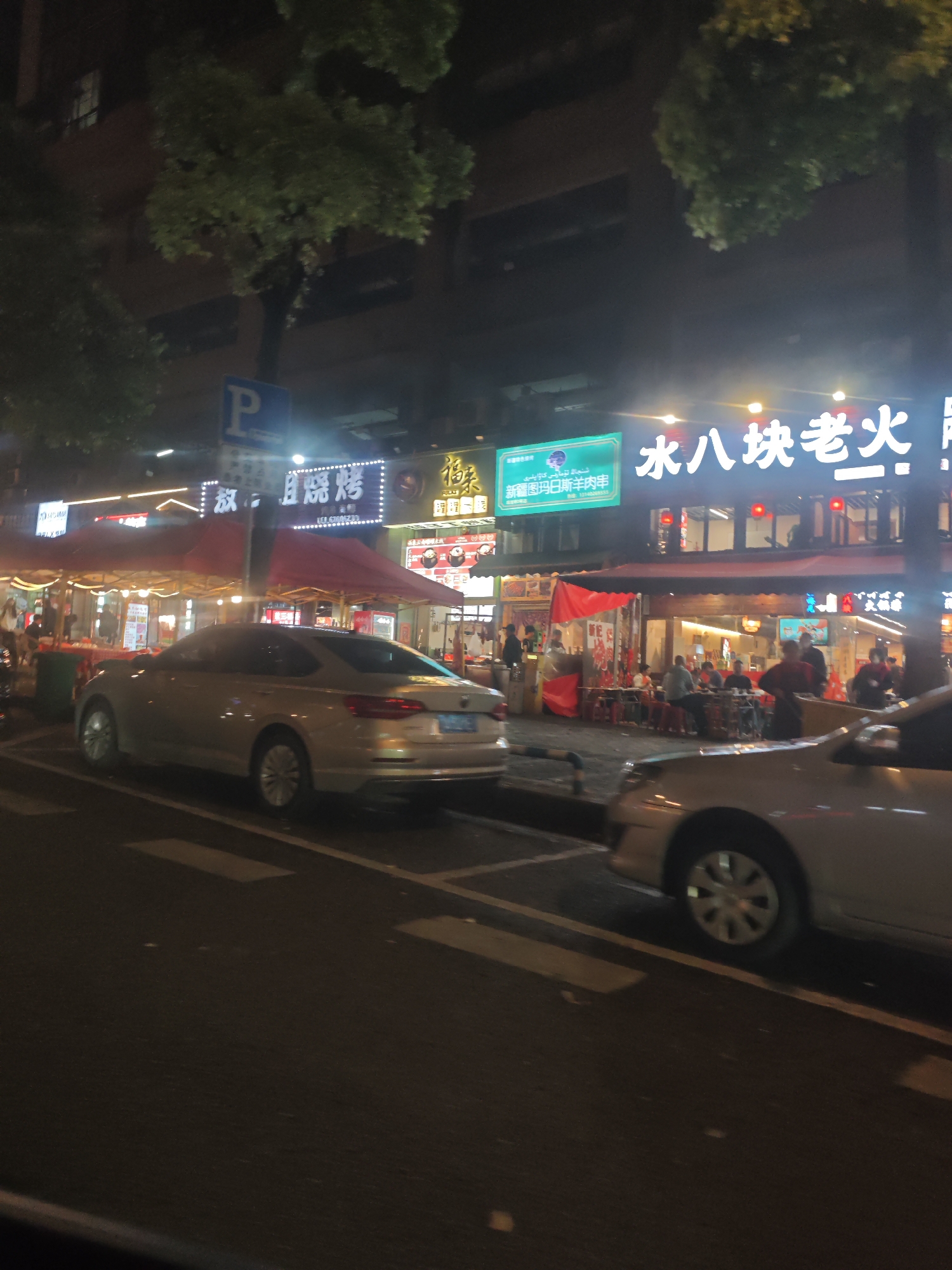 At night in Liangjiang, China, the street scene is bustling with activity. Two cars are parked along the curb, one in front of the other. Above the sidewalk are various outdoor dining establishments, illuminated by string lights and neon signs in Chinese. People are seated at tables, eating and talking. Several individuals are milling about, either customers or passersby. The environment suggests a vibrant nightlife, with restaurants and shops open and active.
