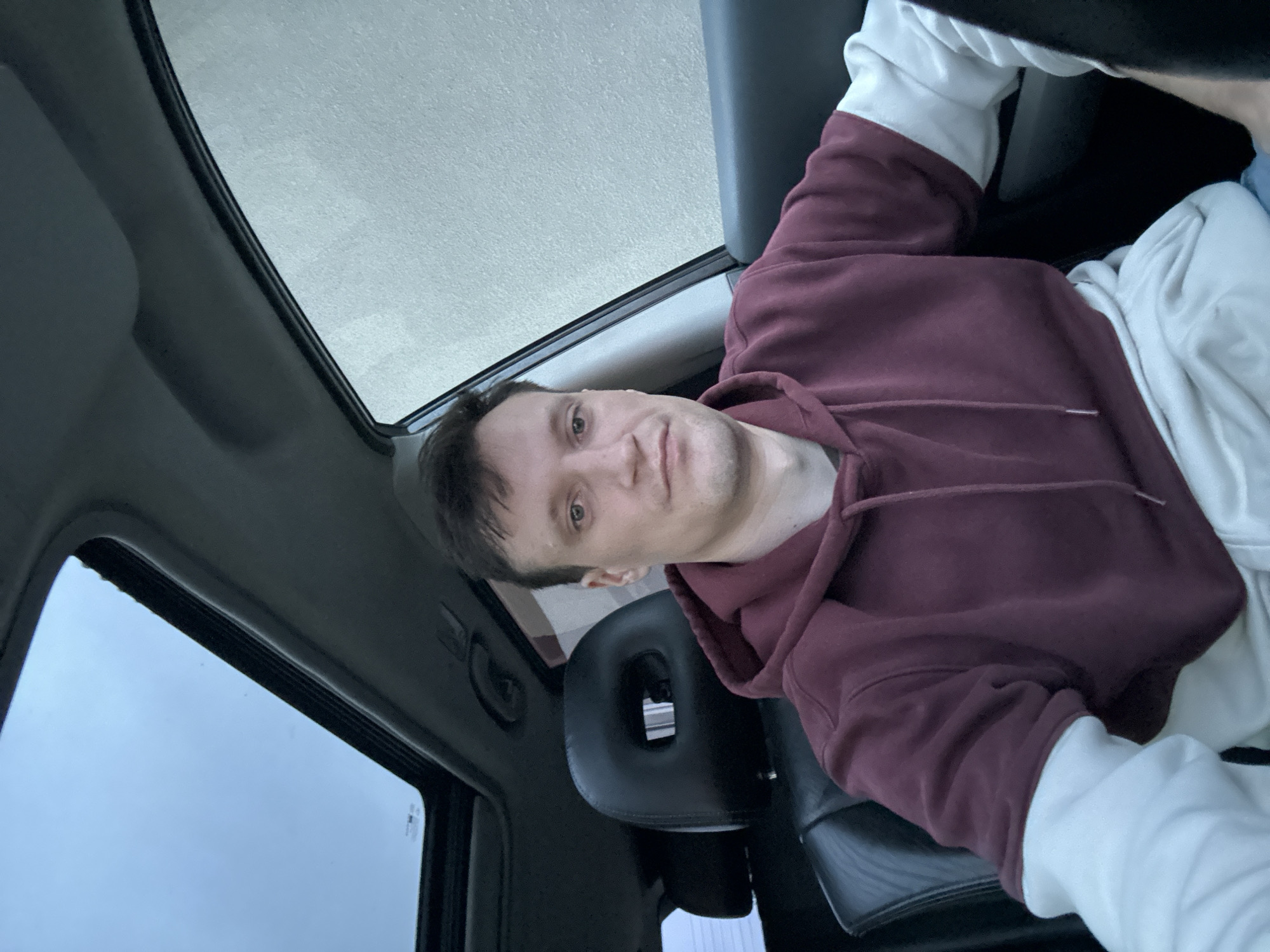 The image shows a medium shot of a man sitting in the driver's seat of a car. He is wearing a maroon hooded sweatshirt layered over a white long-sleeved shirt. The car has a sunroof, which is open, and the interior appears to be dark leather. The background outside the car window is blurry and appears to be a textured wall. The image was taken indoors, likely in a parked car. No specific details about the city of Shatava, Ukraine are visible or inferable from the image.