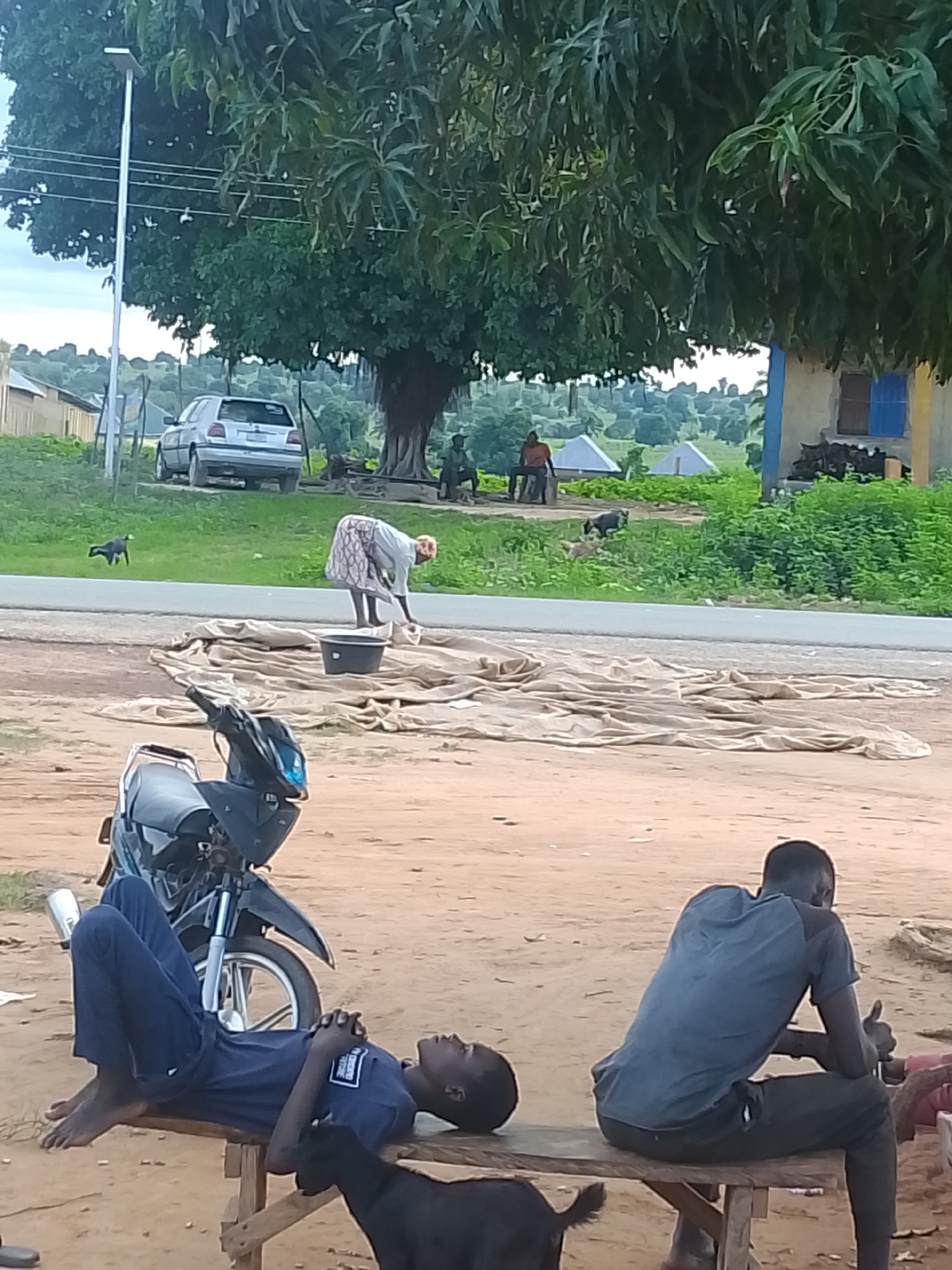 In an outdoor, rural setting, a person wearing a blue uniform is lying on a wooden bench with their legs extended and hands clasped. They are barefoot. A black goat is standing near the bench. Next to the bench is a black and silver motorcycle. Another person, wearing a dark blue shirt and pants, is sitting on a wooden bench to the right of the first person, giving a thumbs-up gesture. A black goat is also near this bench. In the background, a woman in a patterned skirt and light-colored top is bending over, appearing to be working with a bucket and a large piece of fabric spread out on the ground. Further in the background, a silver car is parked near some trees and a building. In the distance, several conical structures are visible in a green, hilly landscape under a cloudy sky. A solar-powered street lamp stands on the left side of the frame.