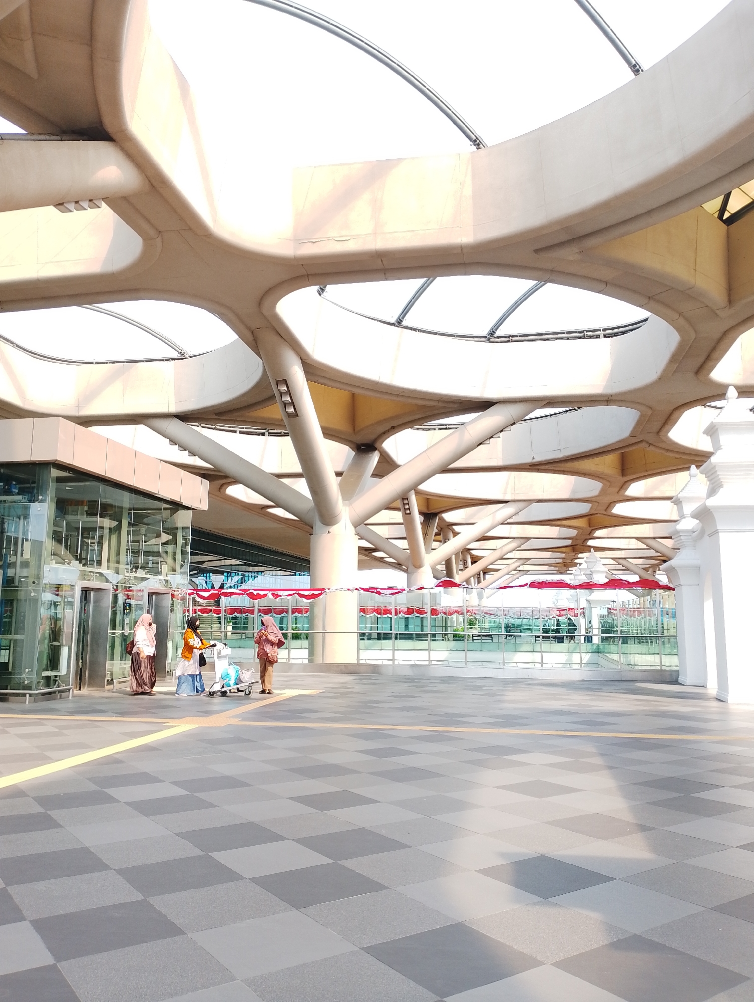 The image depicts the exterior of a modern building at Yogyakarta International Airport (YIA) in Palihan, Indonesia. A group of three individuals, two women in hijabs and one woman in a light orange hijab and blue skirt, are standing on a checkered tiled ground near a glass building entrance. One woman is pushing a luggage cart with blue bags. The building features a distinctive roof structure with large, interconnected circular openings that allow sunlight to filter through. Red and white decorative banners are suspended along a railing in the background. The overall scene suggests a well-maintained, contemporary airport environment bathed in natural light.