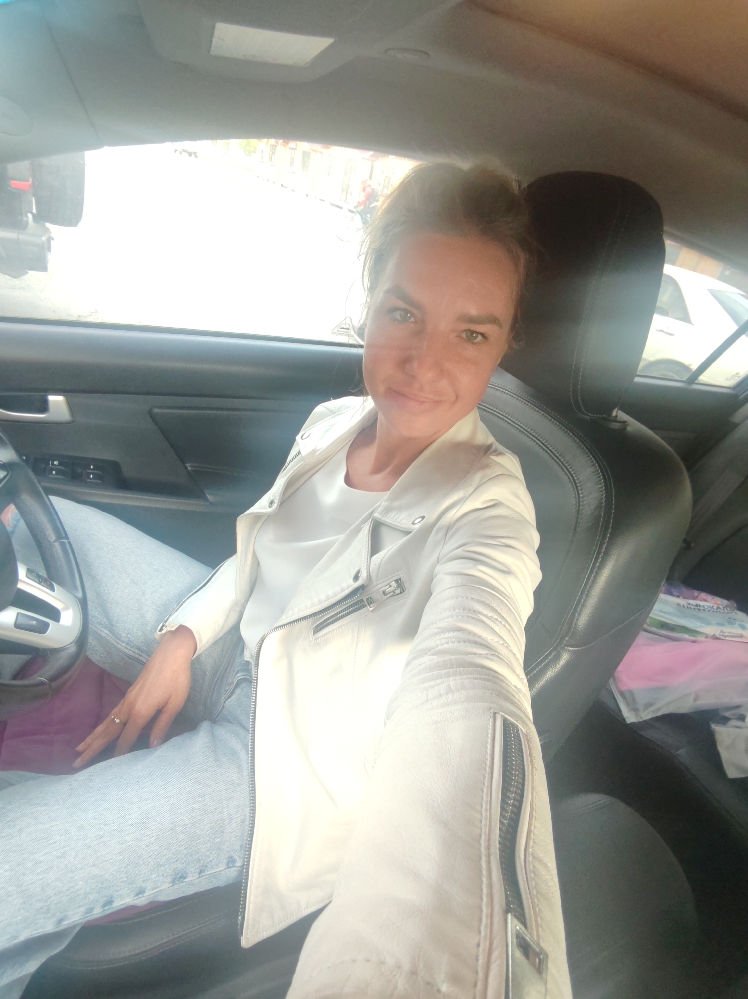 A woman with blonde hair pulled back is taking a selfie in the driver's seat of a car. She is wearing a white leather jacket and light blue jeans. The car's interior is visible, including the steering wheel and car door. Outside the car, buildings and other vehicles can be seen, suggesting an urban environment. The lighting is bright, indicating it is daytime. The location is the city of Petropavlovsk-Kamchatsky, Russia.