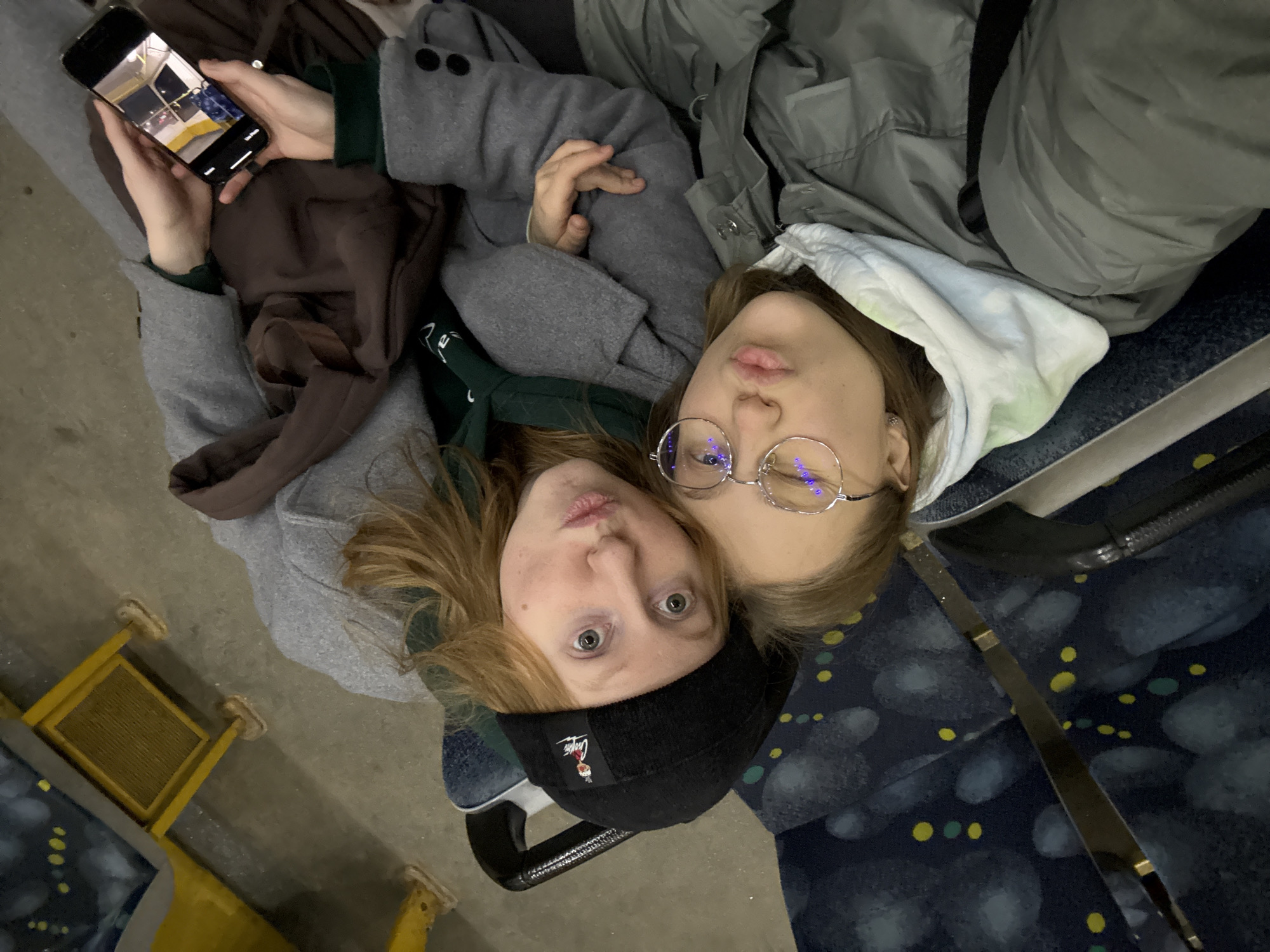 Two young women are lying down on seats in what appears to be a bus or train. One woman is wearing a black beanie and the other has glasses. One is holding a phone. The location is a public transportation vehicle, possibly in Perm, Russia, based on the image on the phone screen. The women are oriented in a way to have their faces in the photo together.