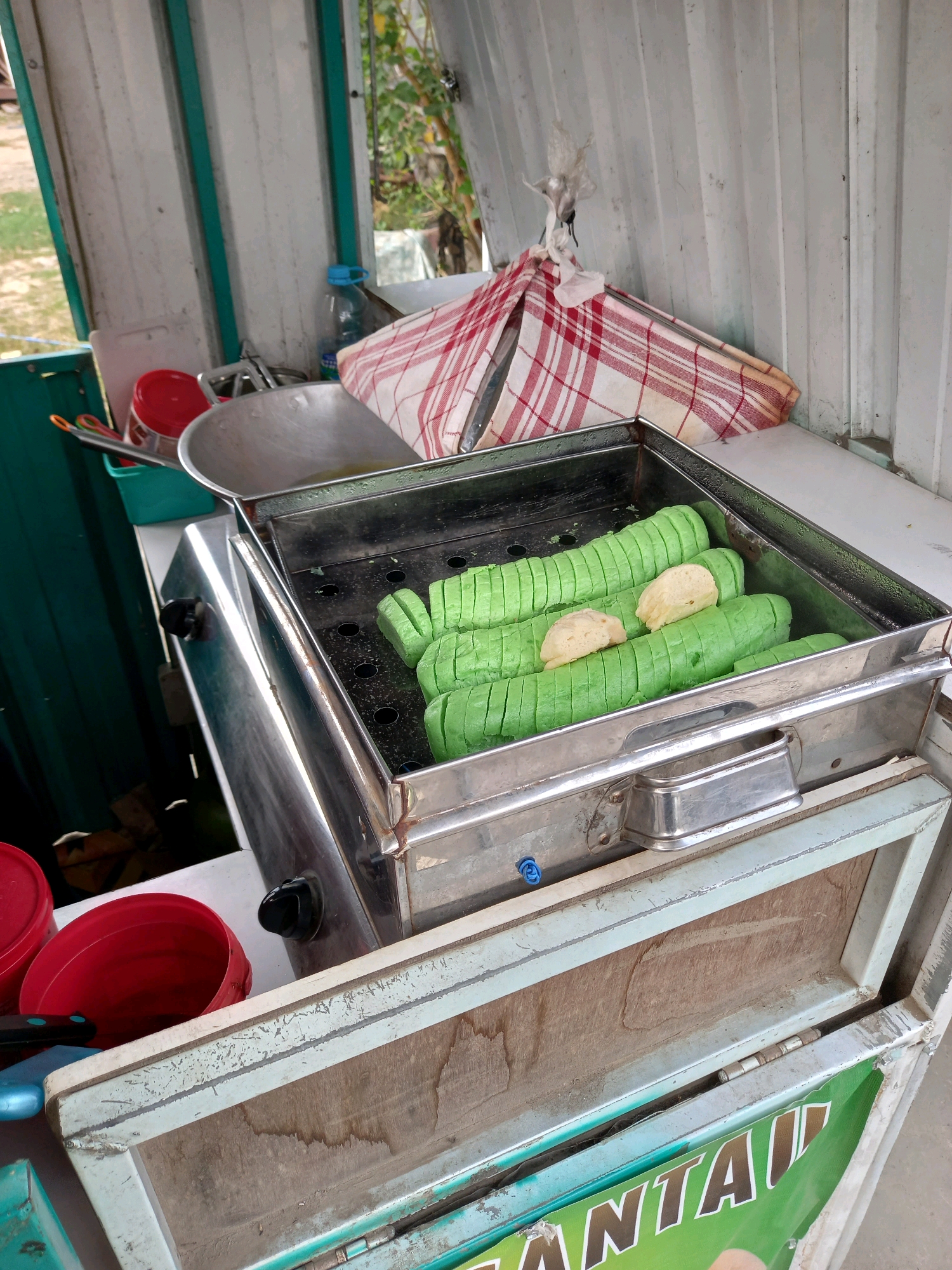 A close-up, eye-level shot captures a stainless steel steamer filled with vibrant green, cylindrical food items that appear to be steamed rolls, cut into bite-sized pieces. Two lighter-colored, soft-looking pieces are placed atop the green rolls. The steamer is positioned on a light-colored, weathered wooden surface, possibly a food cart. The background features a white corrugated metal structure with teal accents, suggesting an outdoor stall or a makeshift cooking area. A red and white checkered cloth is draped over the top edge of the steamer, partially obscuring the metal frame. To the left, a metal wok with a long handle is visible. A blue water bottle and other containers are also present, hinting at a food preparation environment. The overall scene is indicative of a street food vendor setup, likely in a public area within Samarinda, Indonesia, given the context of the provided information about the city. The lighting is natural, suggesting daylight.