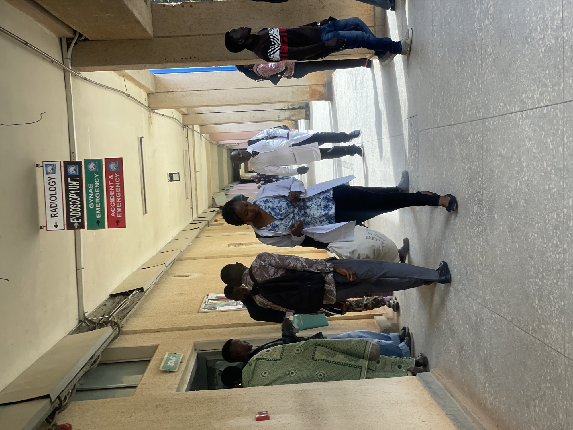 In Gwong, Nigeria, a hospital hallway is depicted, with signs indicating "Radiology," "Endoscopy Unit," "Gynae Emergency," and "Accident & Emergency." Several individuals are present, some appearing to be patients or visitors, and at least two are wearing white coats, suggesting they are medical personnel. One person in a white coat is walking towards the camera, while another is seen further down the hallway. People are dressed in casual attire, including patterned robes, shirts, and jeans. The hallway features a textured beige wall and a concrete floor. Overhead, electrical conduits and lighting fixtures are visible. The atmosphere appears to be that of a functional, though possibly busy, medical facility.