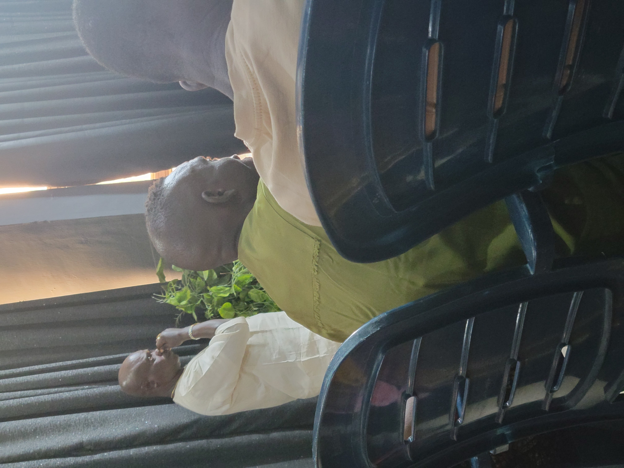 In Aba, Nigeria, a dimly lit indoor scene shows three individuals. The person on the left, a bald man wearing a light-colored outfit, stands with his hand to his mouth, looking downwards. In the center, a man with a bald head and a dark green garment is seated, facing away from the camera. To the right, another individual, whose back is to the camera and is wearing a light-colored shirt, is also seated. They appear to be in a room with dark curtains and a thin beam of light entering from the right. The foreground is dominated by the backs of dark plastic chairs.