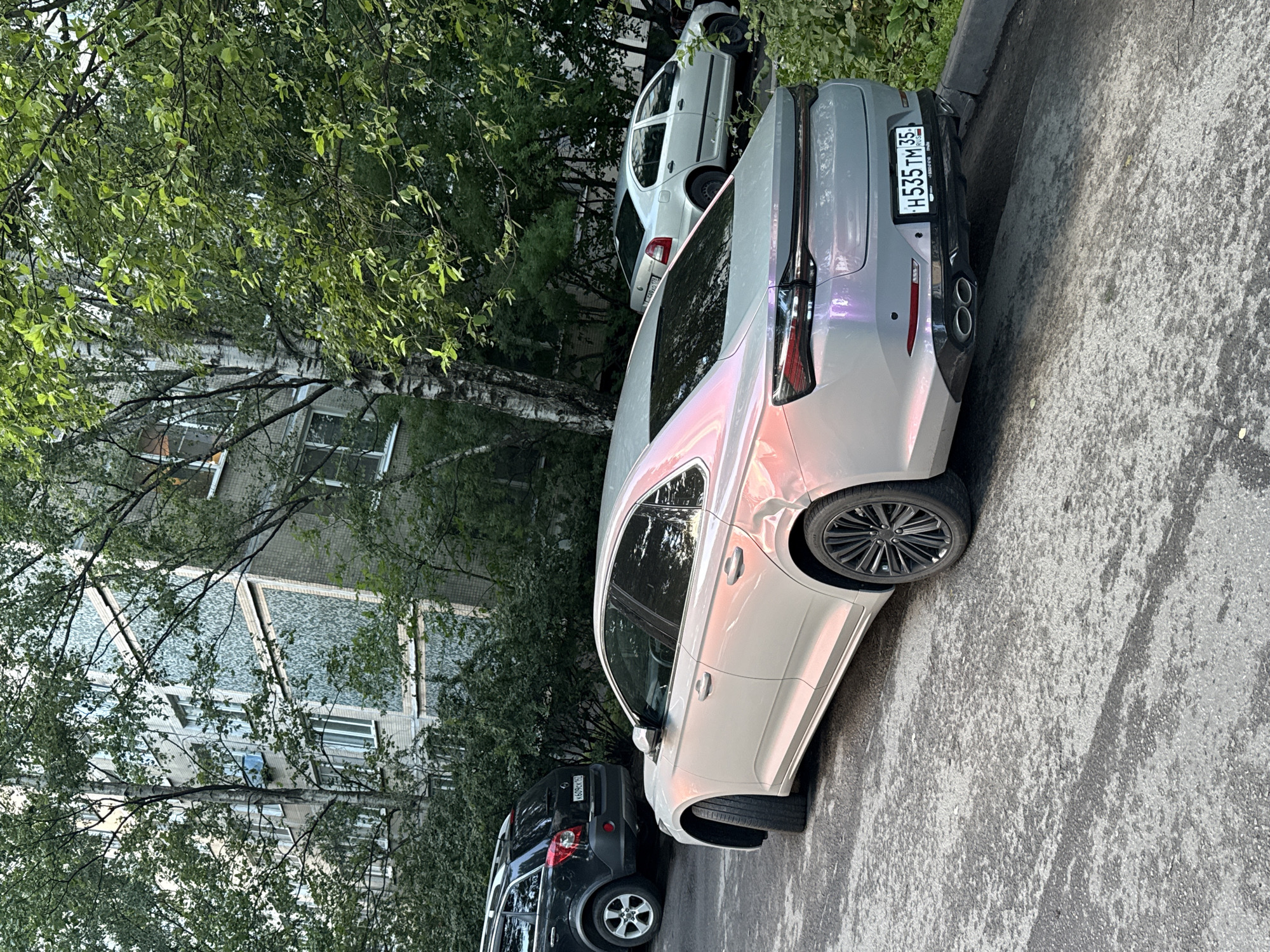 A light-colored sedan with a pearlescent finish is parked on an asphalt surface in front of a multi-story apartment building in St. Petersburg, Russia. The car has a license plate that reads "H535TM 35 RUS". To the left of the sedan, a dark-colored SUV is parked. The background is filled with lush green trees and the facade of a typical Soviet-era apartment building with numerous windows. The scene appears to be a residential area within the city.