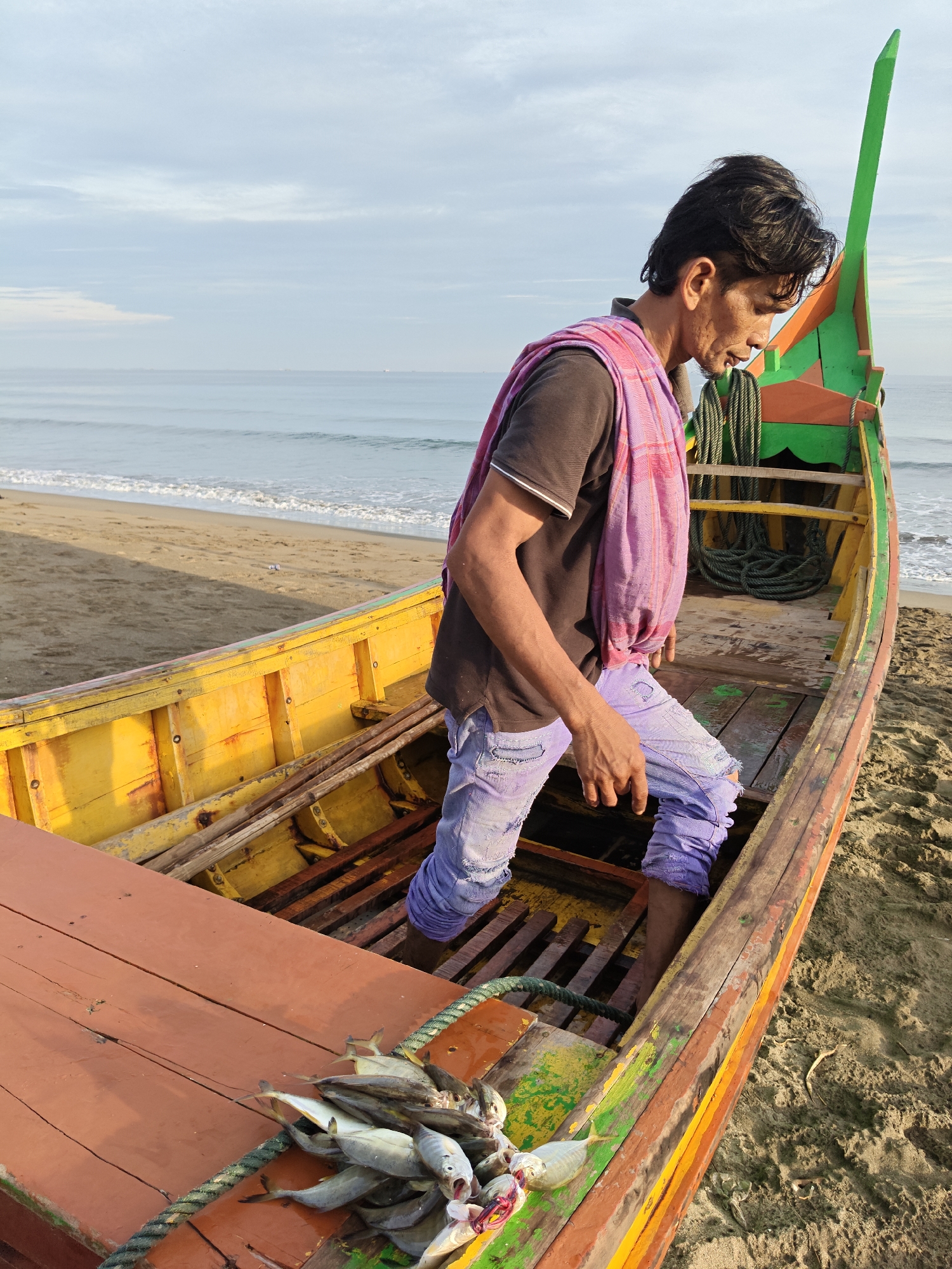 In Curehtunong, Indonesia, an individual is standing in a colorful wooden fishing boat docked on a sandy beach. The boat is predominantly yellow with red and green accents, and a vibrant green decorative element at its bow. The person, dressed in a dark t-shirt, purple distressed jeans, and a pink and purple checkered scarf draped over their shoulders, is looking down into the boat. A pile of freshly caught fish rests on the wooden deck in the foreground of the boat. The background consists of a calm ocean with a gently receding shoreline and a sky with scattered clouds.