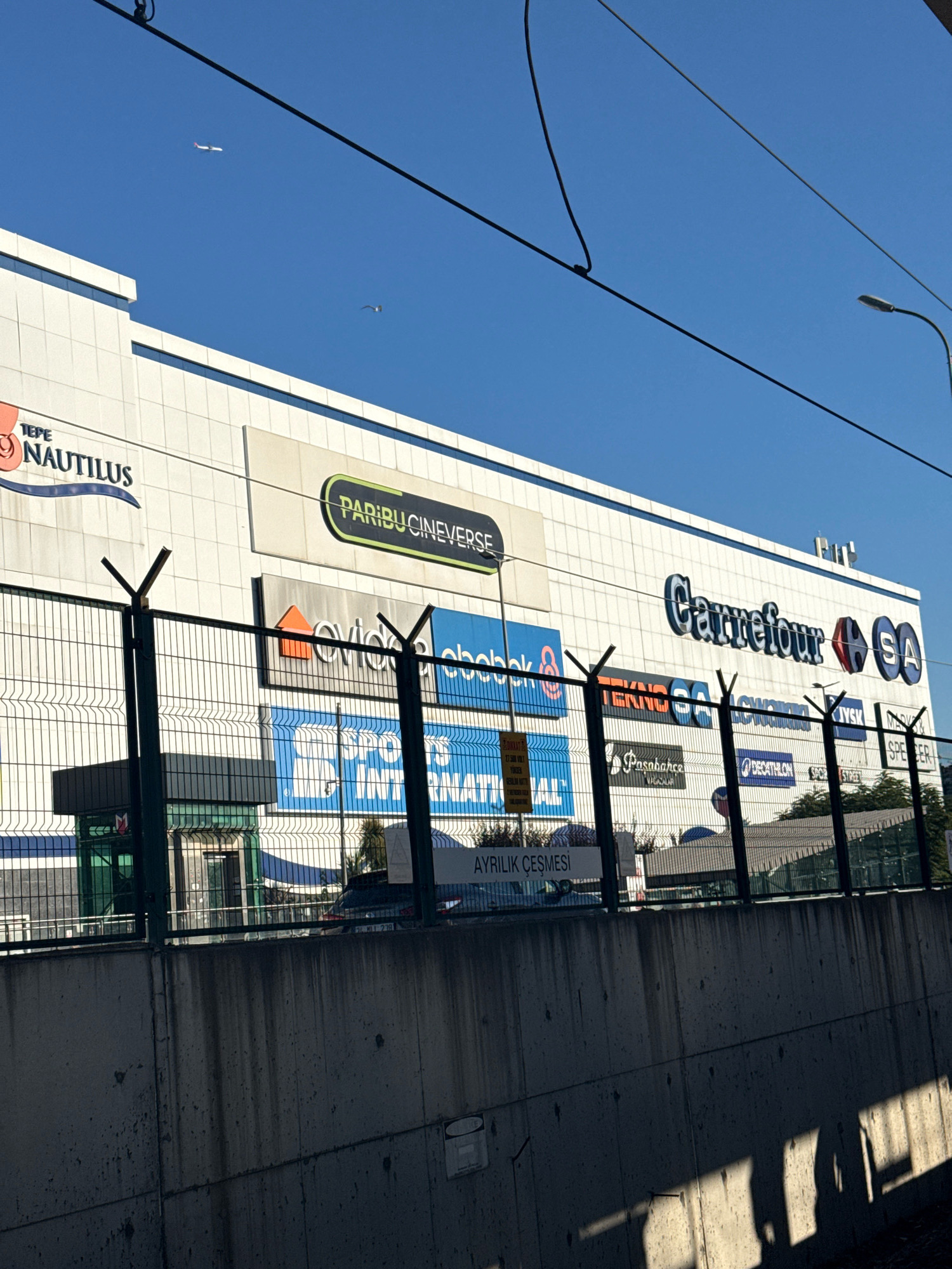 The image captures a bright, sunny day in a city, with a large, modern shopping mall as the central focus. The mall's facade is white with prominent signage, including "TEPE NAUTILUS", "PARIBUCINEVERSE", and "Carrefour SA". Various other store logos are visible, such as "evidea", "e-bebek", "Teknosa", "LC Waikiki", "Decathlon", "Paşabahçe", and "Superstep". In the background, a clear blue sky is dotted with two distant white airplanes and a single bird. The mall is enclosed by a tall chain-link fence with metal spikes on top, and a concrete wall runs along the foreground. Several trees are visible behind the fence. The overall impression is one of a commercial hub bathed in natural light.
