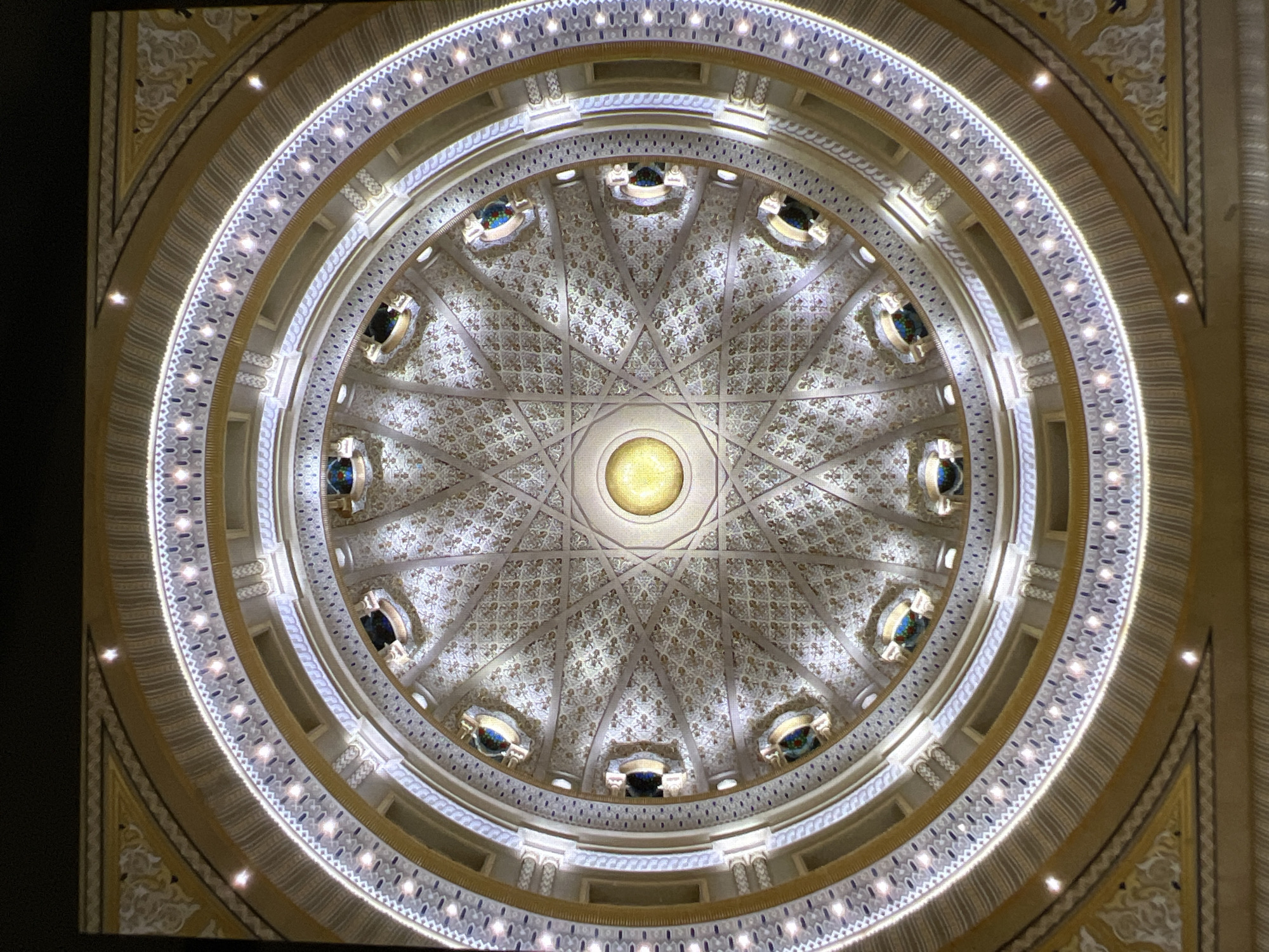 The image shows a low-angle, full-frame shot of an ornate domed ceiling. The ceiling is circular and features a central golden orb surrounded by intricate geometric patterns. The patterns are repeated in a radial design, with radiating lines and detailed tessellations in shades of white and gold. Along the outer edge of the dome, there are evenly spaced illuminated recessed panels with what appear to be small, colorful stained-glass inserts. These panels are set within a decorative border that includes gold trim and a white textured band. The overall impression is one of grandeur and detailed craftsmanship. The image does not contain any people or indicate a specific location within Verona, Italy, or any other city.