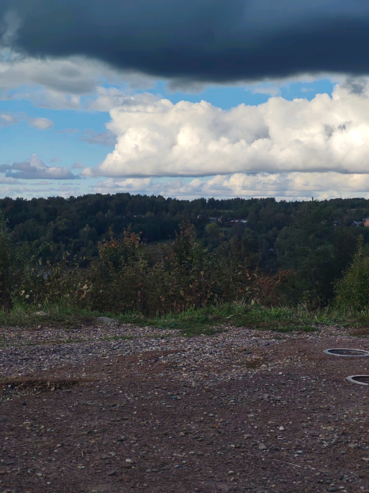 The image depicts a landscape view from a gravelly area in the city of Plyos, Russia. In the foreground, there is a ground covered with small stones and some patches of green grass. Further back, a dense forest stretches across the horizon, with scattered houses visible among the trees. Above the forest, a dramatic sky is filled with a mix of dark, heavy clouds and bright, puffy white clouds, with patches of blue sky showing through. Two round, dark objects, possibly manhole covers or drainage openings, are visible on the gravelly surface in the lower right quadrant of the image.