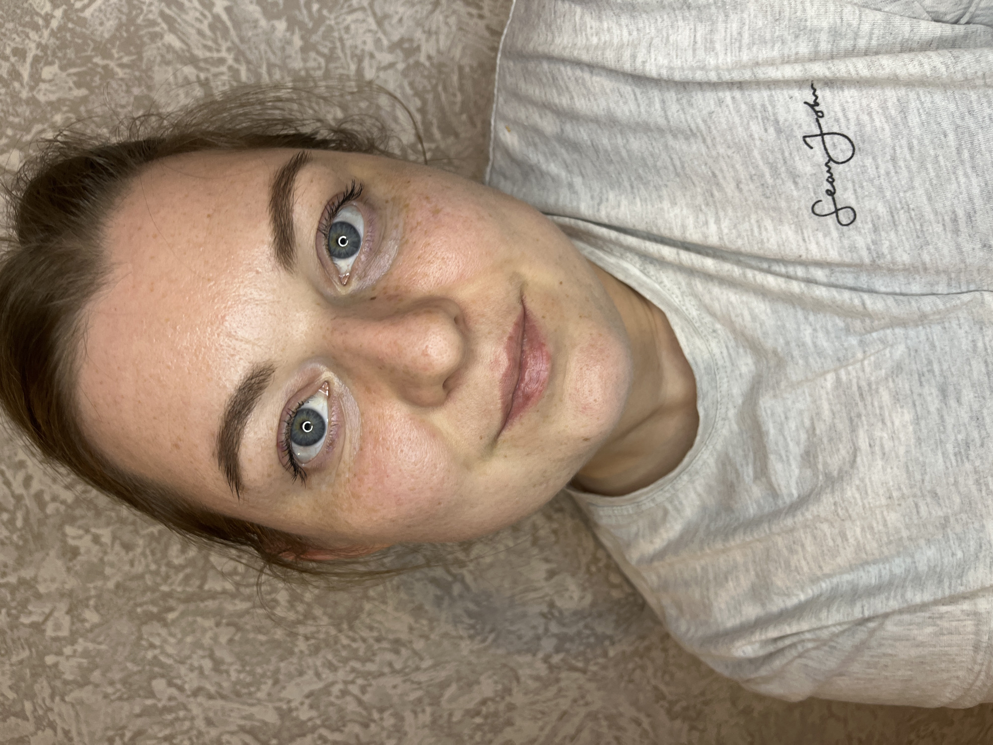 A woman lies at an angle against a patterned surface, likely a bed or couch. She faces the camera with a slight smile. She has fair skin with freckles, blue eyes, and brown hair. She is wearing a gray t-shirt with the words "Sean John" written vertically on the upper right side of the shirt. The image does not contain information to ascertain a specific location in Раздолье, Russia.